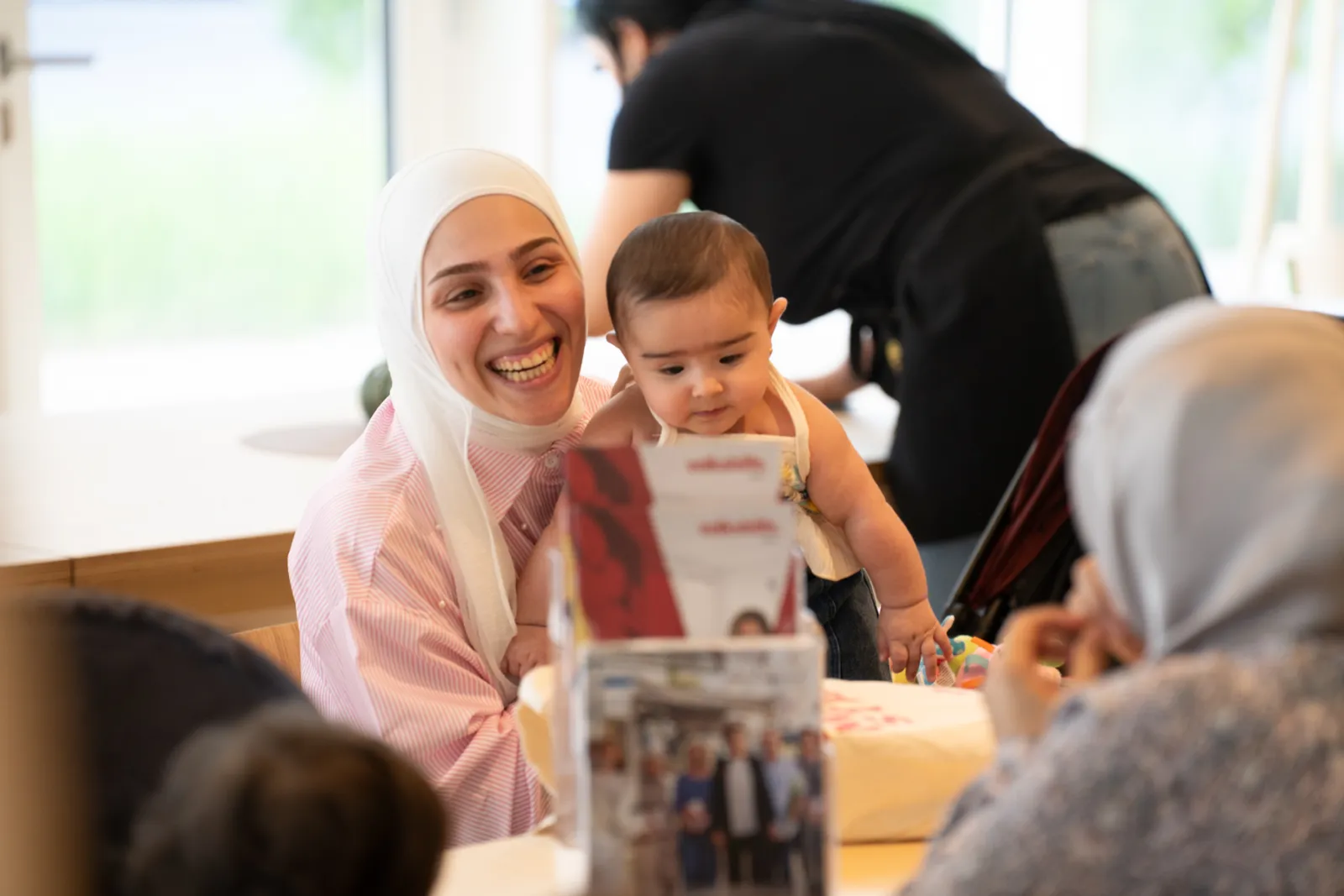 Frau im weißen Hijab hält ein Baby und lächelt; sie sitzt an einem Tisch in hellem Gemeinschaftsraum.
