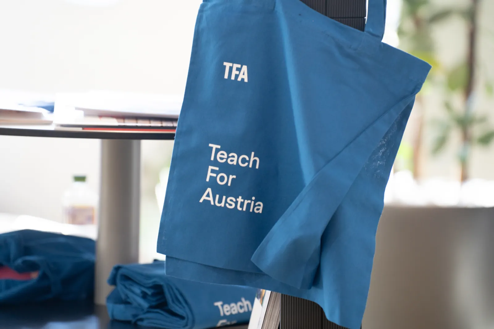Blaue Tragetasche mit Aufschrift Teach For Austria hängt an einem Tisch im Innenraum.