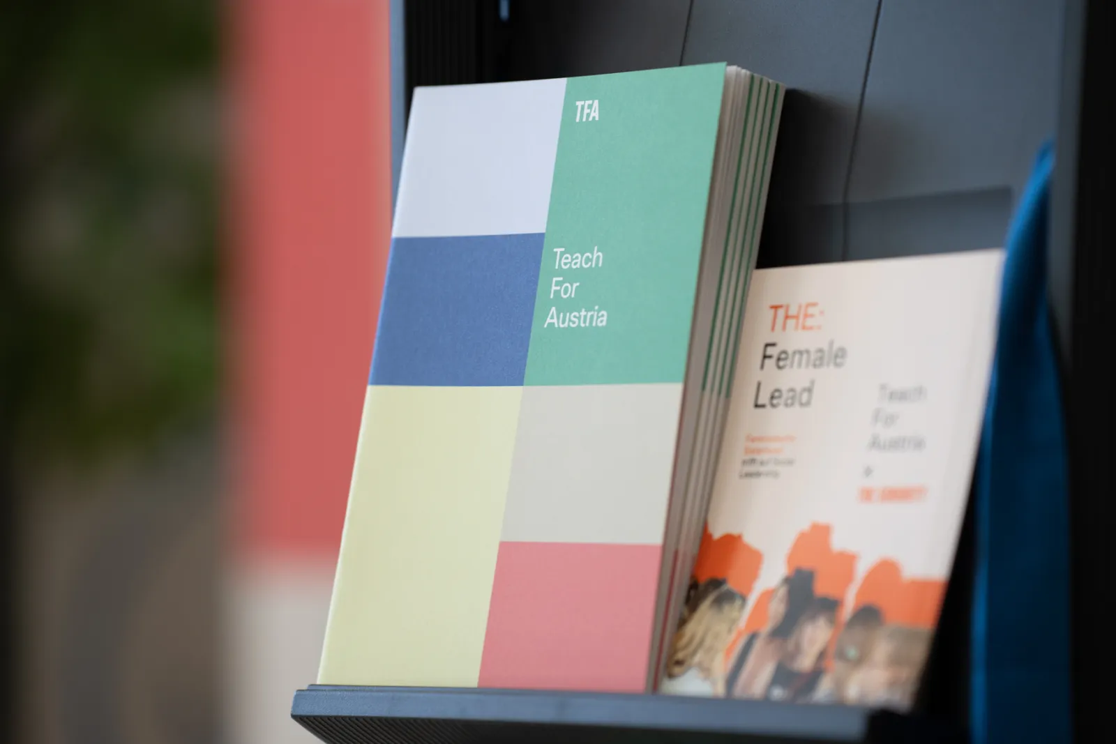 Bunte Broschüren auf einem Regalständer, Titel Teach For Austria und THE: Female Lead sichtbar.