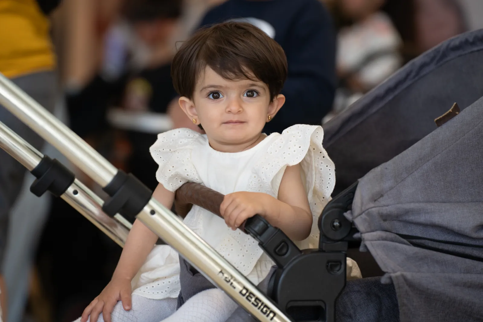 Kleinkind im weißen Spitzenkleid sitzt im Kinderwagen, hält Griff, schaut zur Kamera; unscharfer Hintergrund.