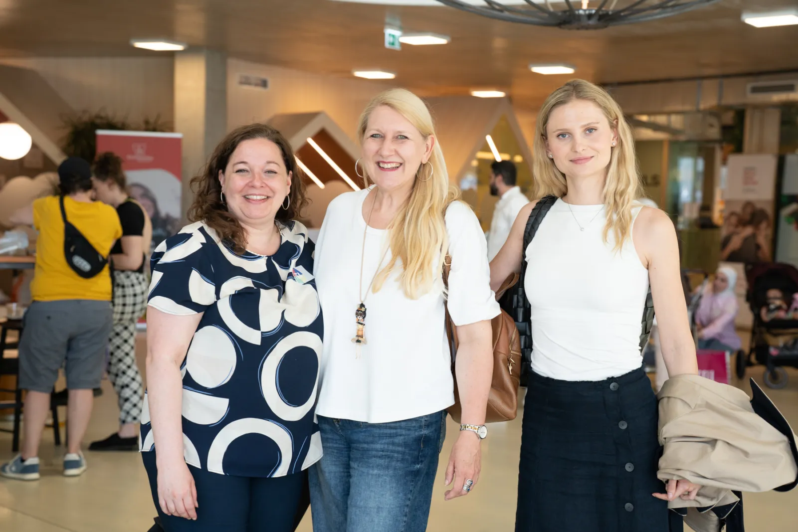 Drei Frauen posieren lachend in hellem Innenraum einer Veranstaltung; Hintergrund mit Ständen und Besuchern.