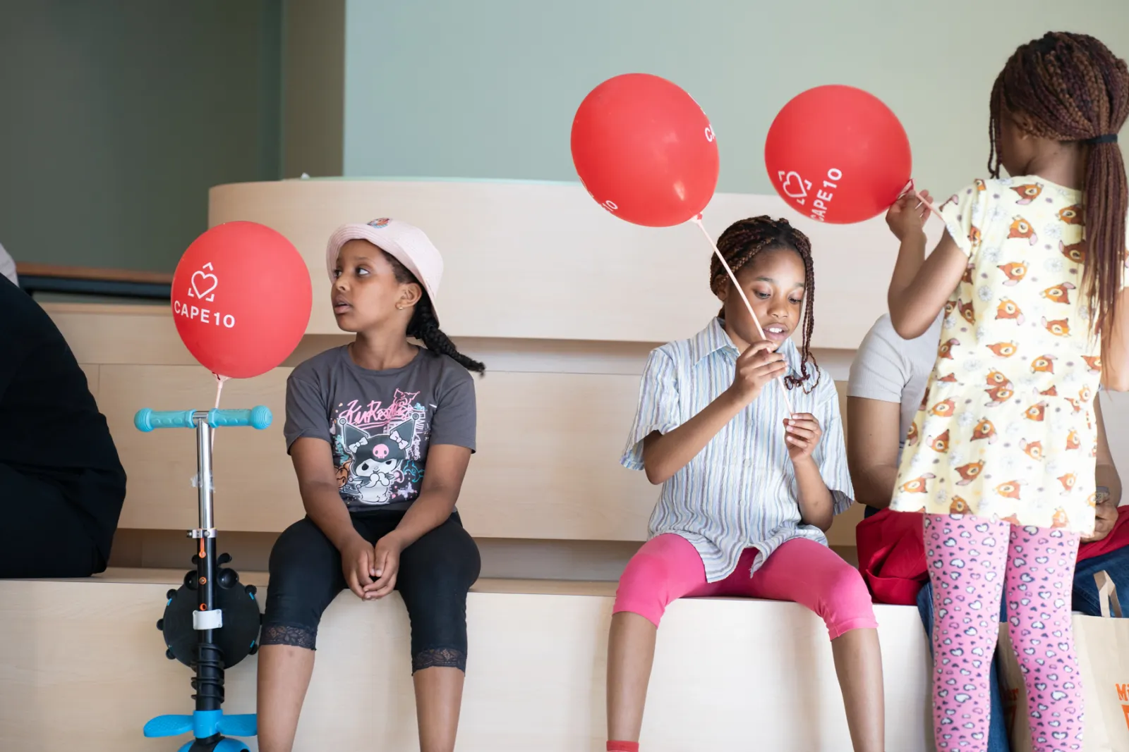 Drei Kinder im Wartebereich auf einer Holzbank; mittleres Kind hält roten Ballon, links sitzt Kind mit Mütze, rechts steht Kind in gemustertem Kleid neben Scooter.
