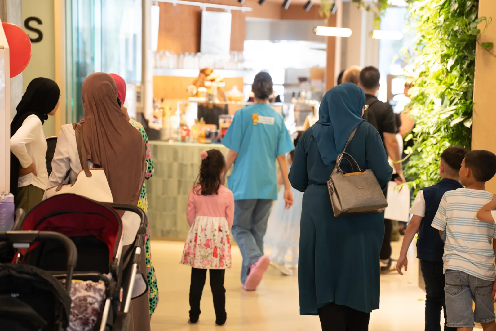 Person mit blauem Hijab und grauer Handtasche geht durch Einkaufszentrum zum Café; weitere Passanten und ein Kind im Hintergrund.