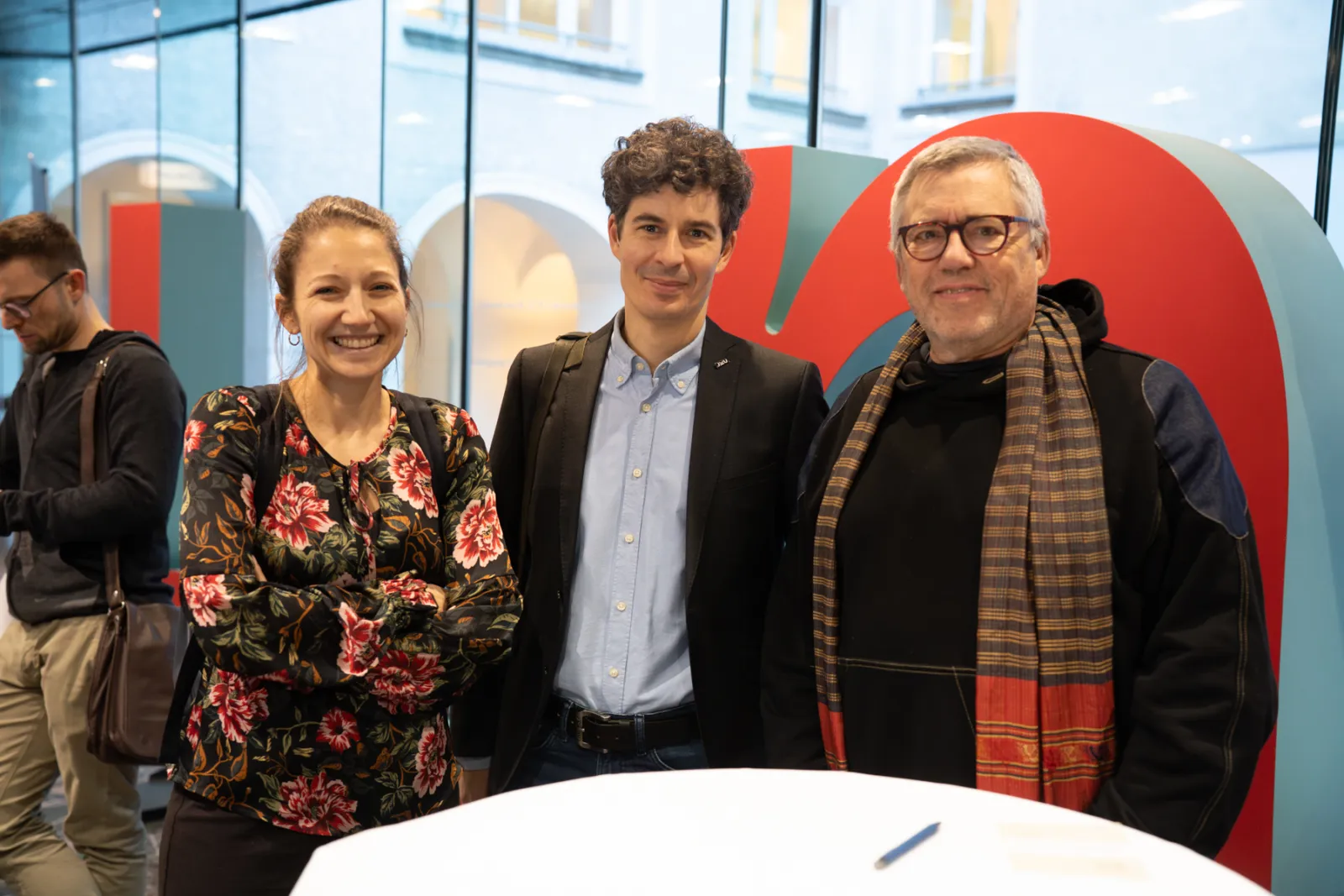 Drei Personen posieren lächelnd vor farbiger Kunstinstallation in Innenraum; links Person in floralem Oberteil, Mitte Person im Blazer, rechts Person mit Schal.