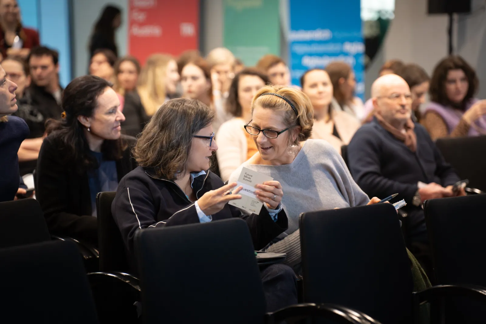 Zwei Frauen im Vordergrund diskutieren über ein Blatt Papier in einem Konferenzsaal, hinter ihnen sitzende Zuschauer.