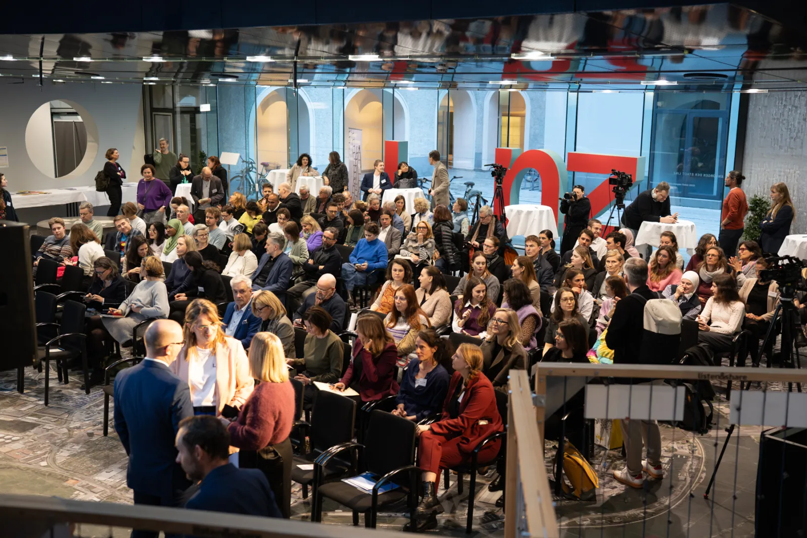 Große Zuschauermenge in moderner Konferenzhalle; Kamerateams filmen, im Vordergrund diskutieren zwei Personen.