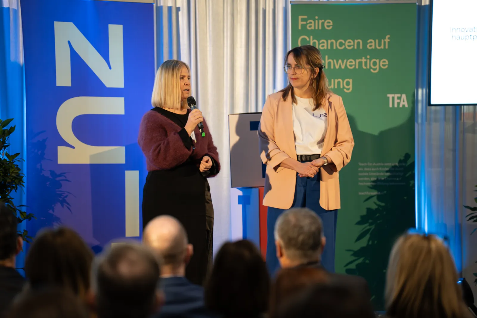 Zwei Personen auf einer Veranstaltungsbühne vor Publikum; eine Person spricht ins Mikrofon, die andere schaut zu.