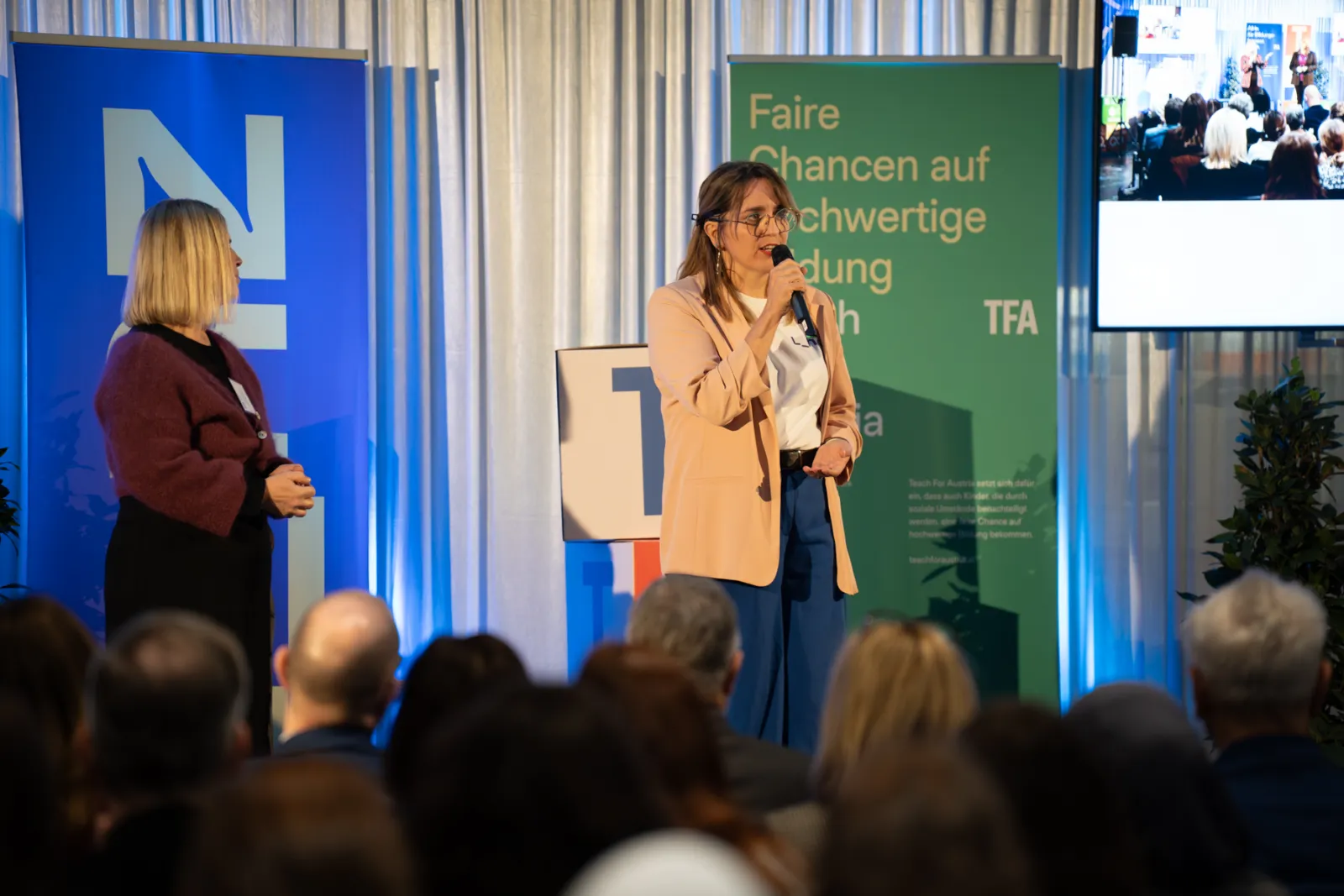 Zwei Personen auf einer Konferenzbühne; eine Person spricht ins Mikrofon, die andere hört zu; Hintergrund mit blauen und grünen Bannern.