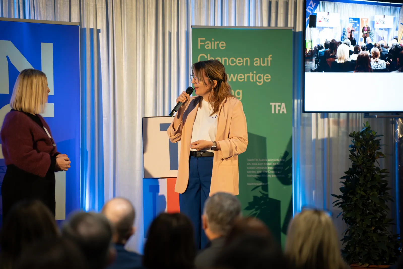Frau mit Mikrofon spricht auf Konferenzbühne; zweite Frau hört zu. Grünes Banner mit 'Faire Chancen' im Hintergrund, Monitor zeigt Publikum.