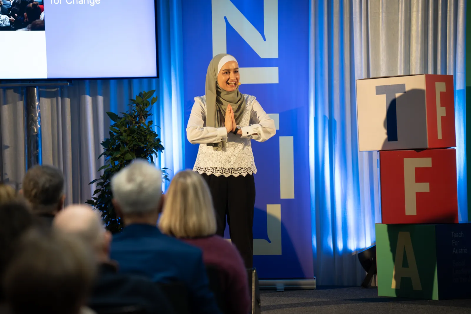 Frau im Hijab steht auf Bühne und begrüßt das Publikum bei einer Konferenz; im Hintergrund blaues Bühnenbanner, rechts bunte Buchstabenwürfel.