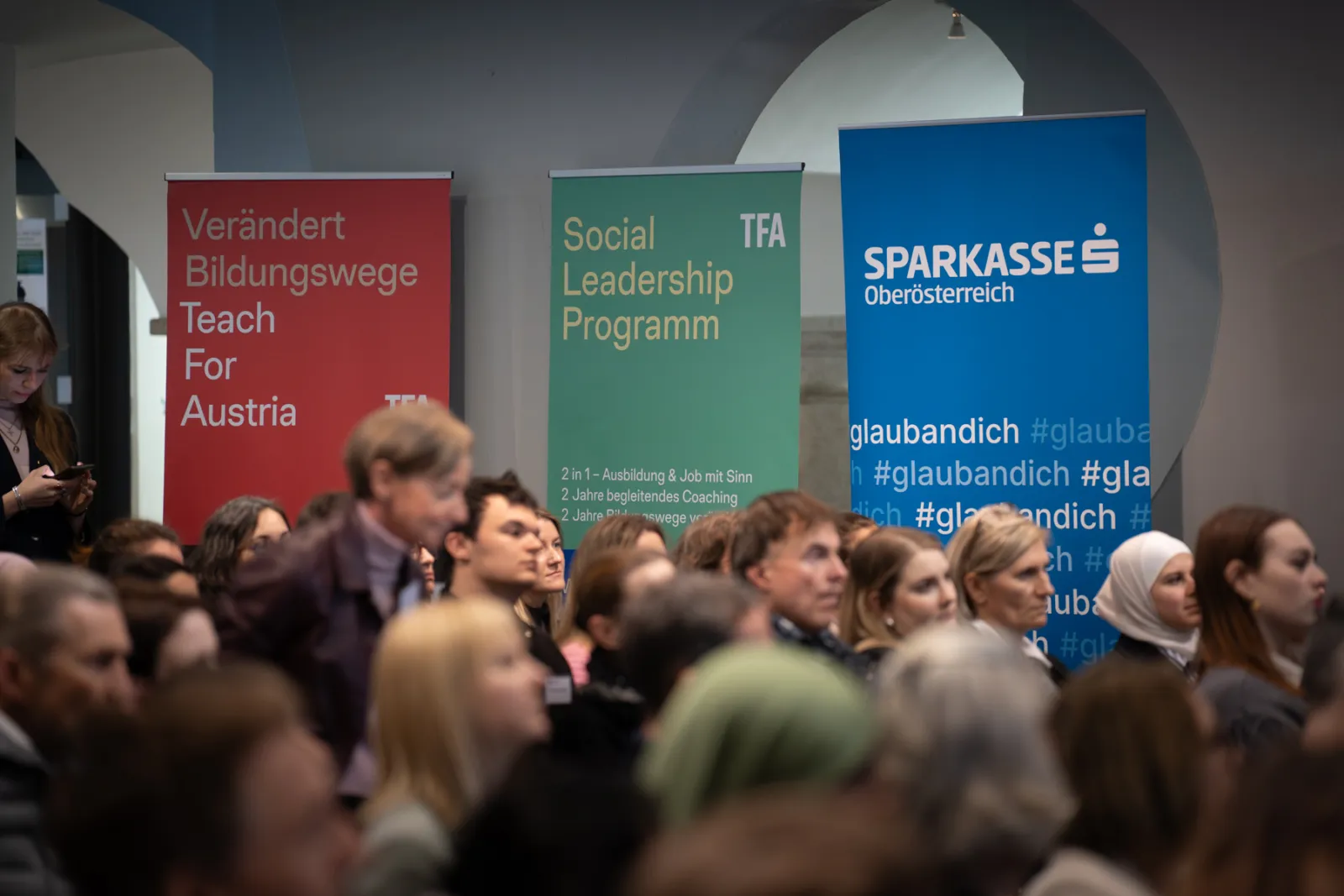 Publikum lauscht einer Konferenz in einem Veranstaltungsraum; im Hintergrund rote, grüne und blaue Banner.