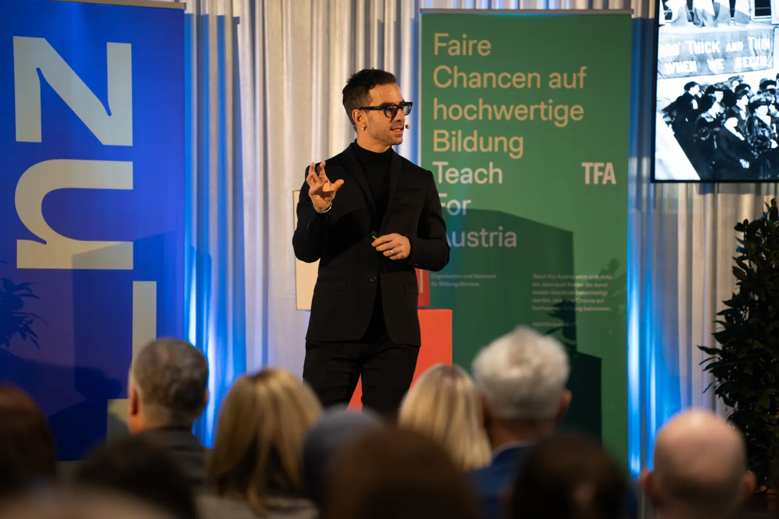 Mann in schwarzem Anzug mit Brille hält Vortrag auf der Bühne; grünes Banner mit Faire Chancen auf hochwertige Bildung Teach For Austria; Publikum.