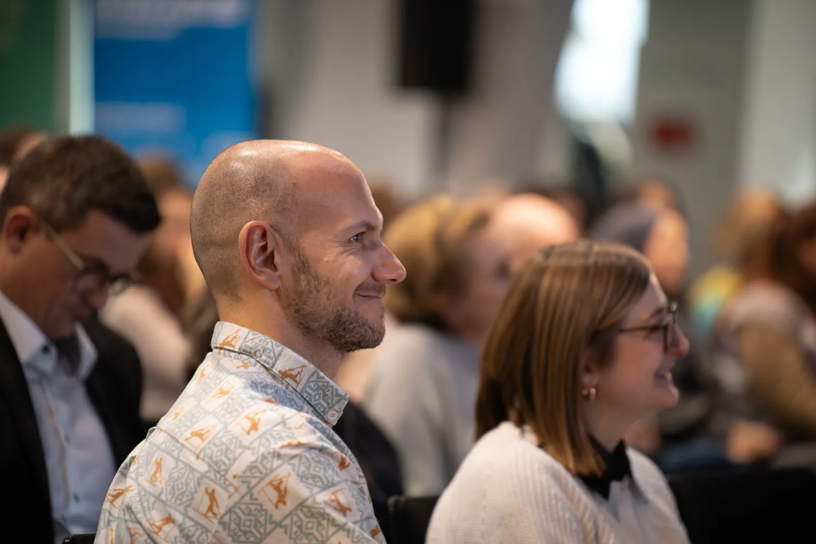 Person mit Glatze lächelt im Vordergrund bei einer Konferenz.