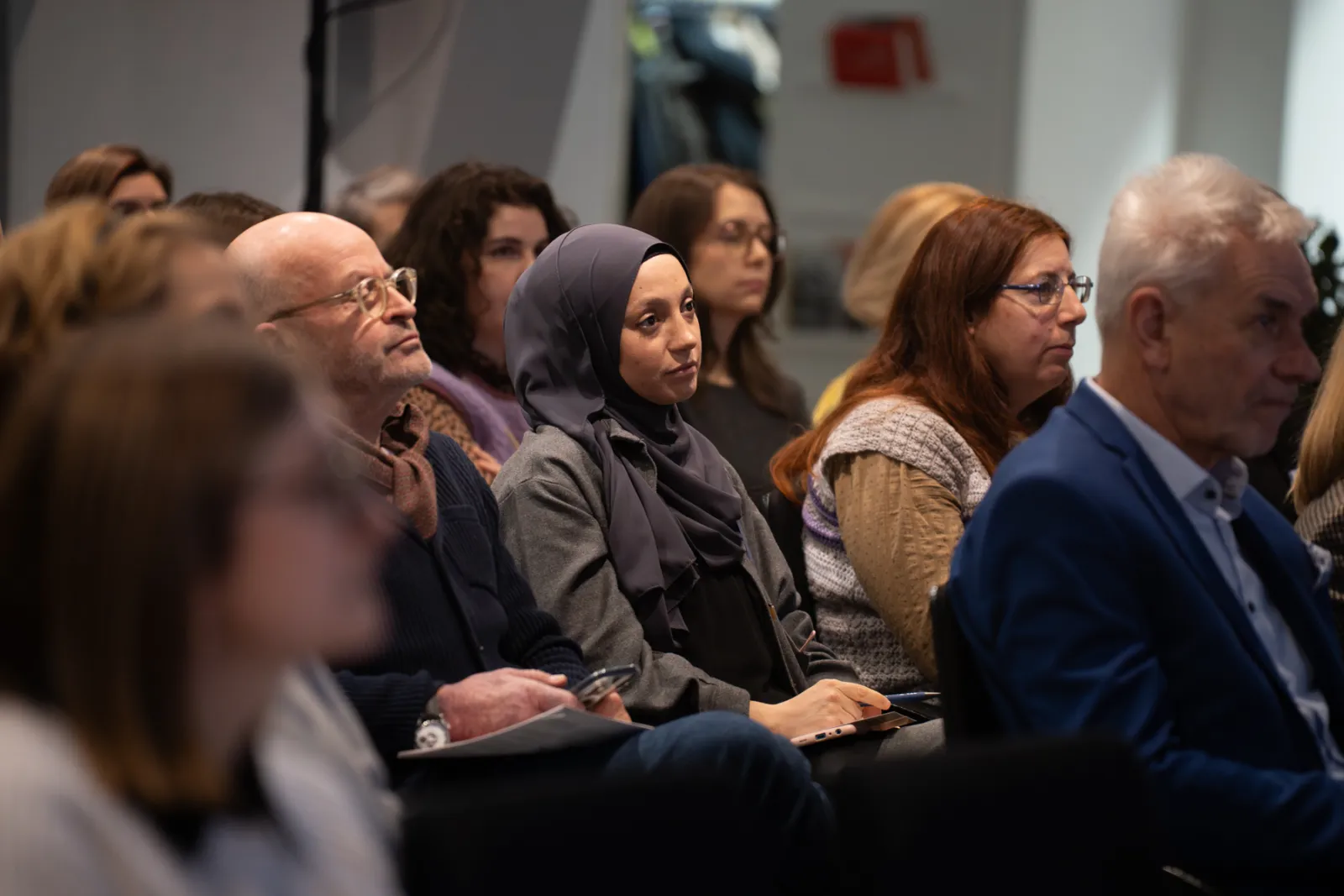 Zuhörergruppe im Konferenzsaal; zentrale Frau mit dunkelgrauem Hijab hört aufmerksam zu, umgeben von weiteren Teilnehmern.