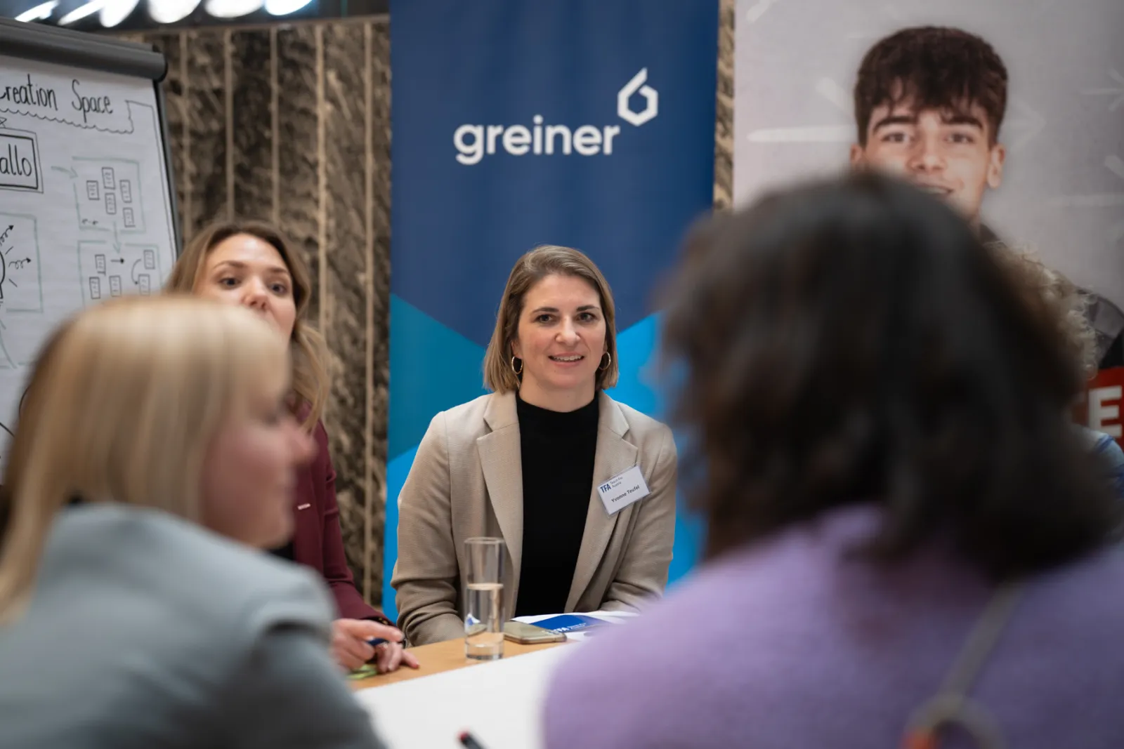 Person in beigem Blazer sitzt am Messestand-Tisch; lächelt, Banner mit 'Greiner' im Hintergrund.