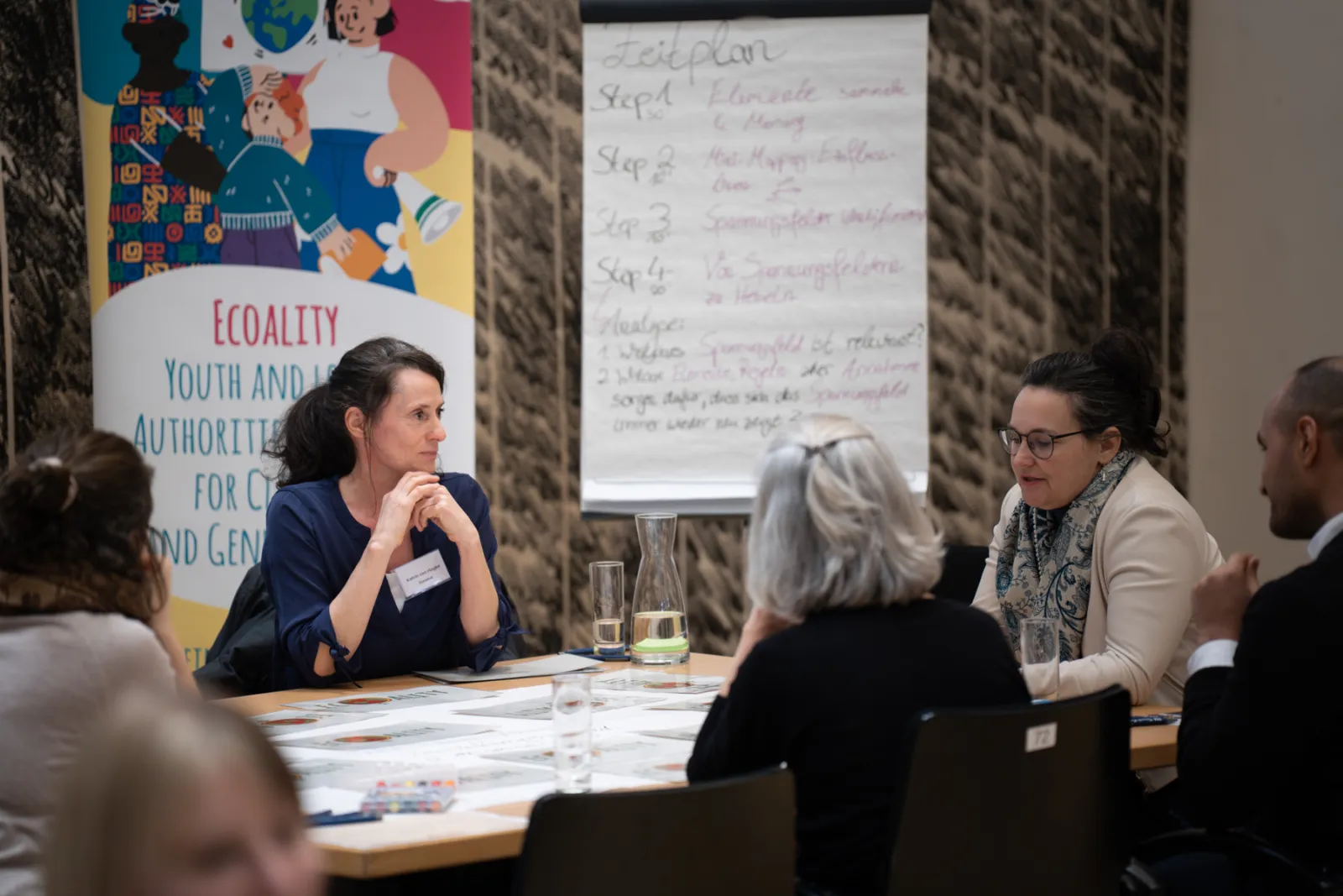 Gruppe von Erwachsenen diskutiert an Konferenztisch; Seminarraum mit Flipchart-Notizen und buntem Banner im Hintergrund.