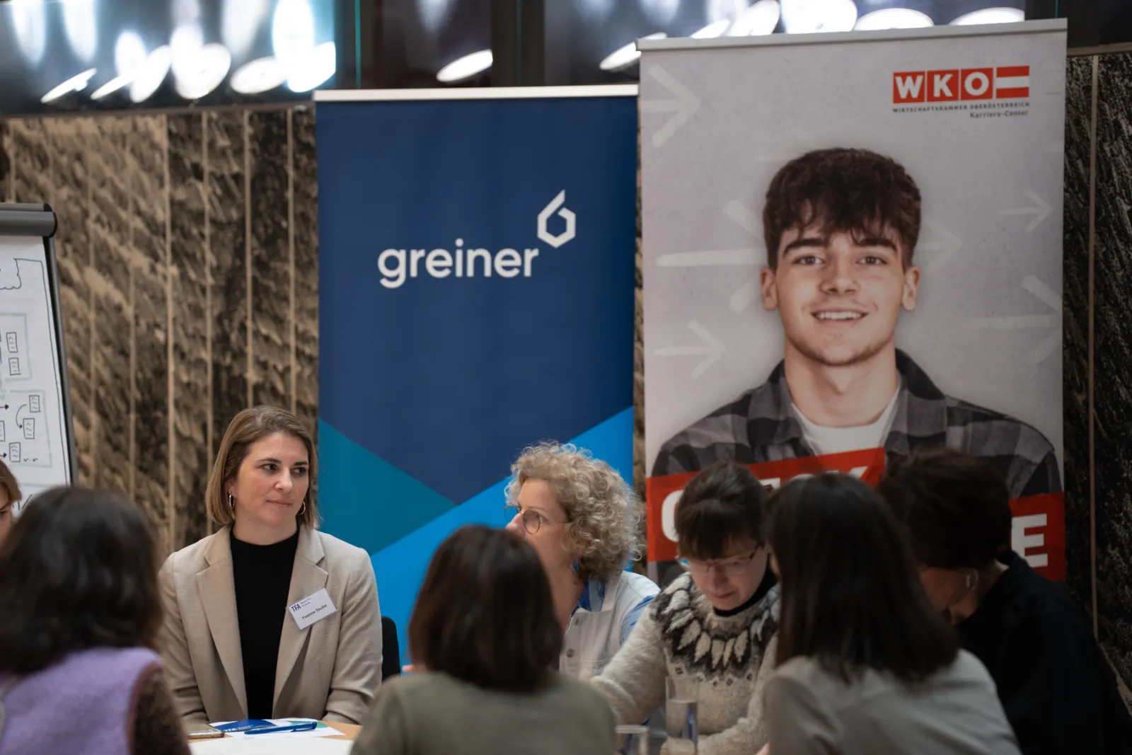 Personengruppe sitzt an einem Tisch bei einer Karriereveranstaltung; Banner im Hintergrund.