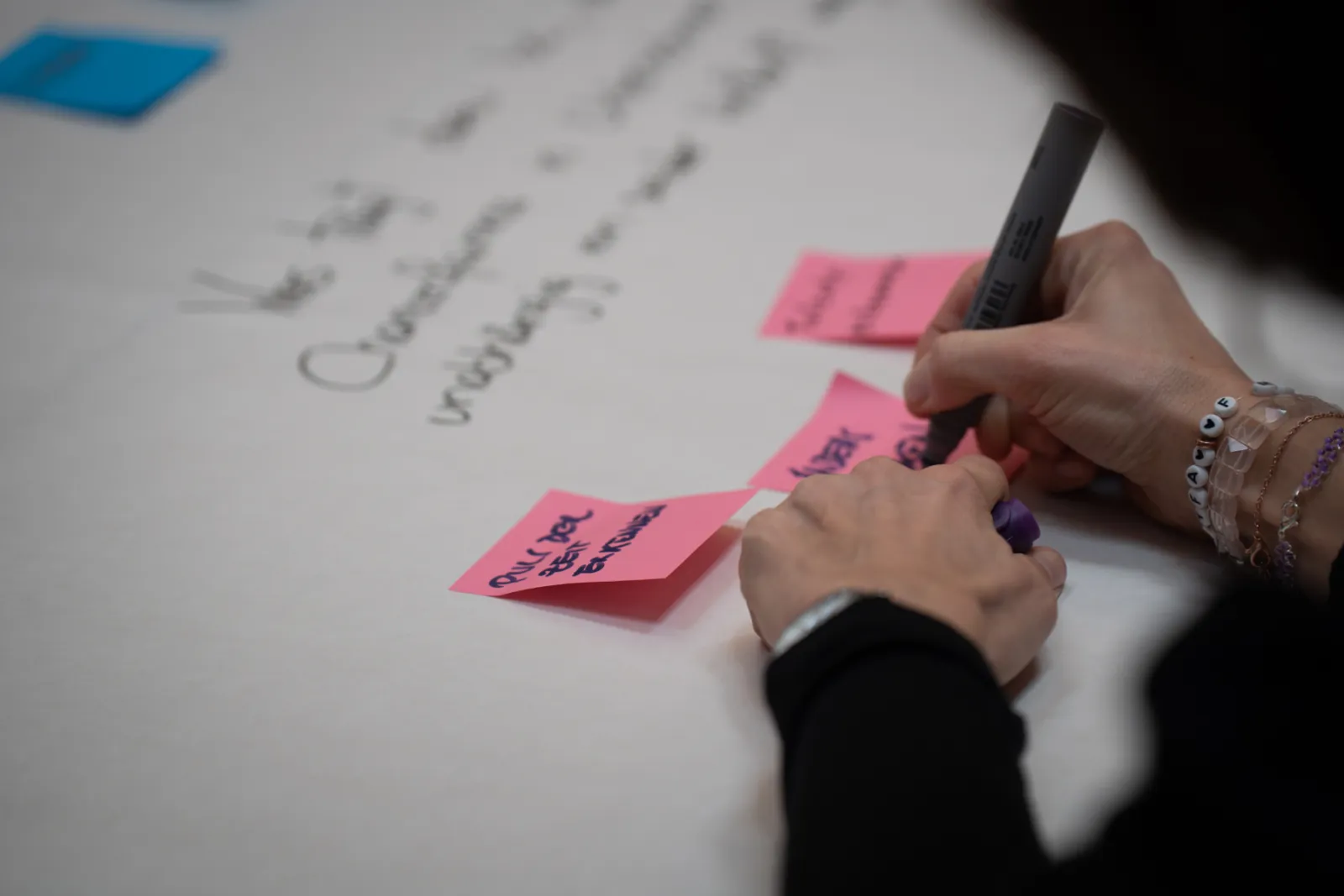 Nahaufnahme von Händen, die mit Marker auf rosa Post-it-Notizen auf einem weißen Tisch schreiben; Armbänder am Handgelenk.