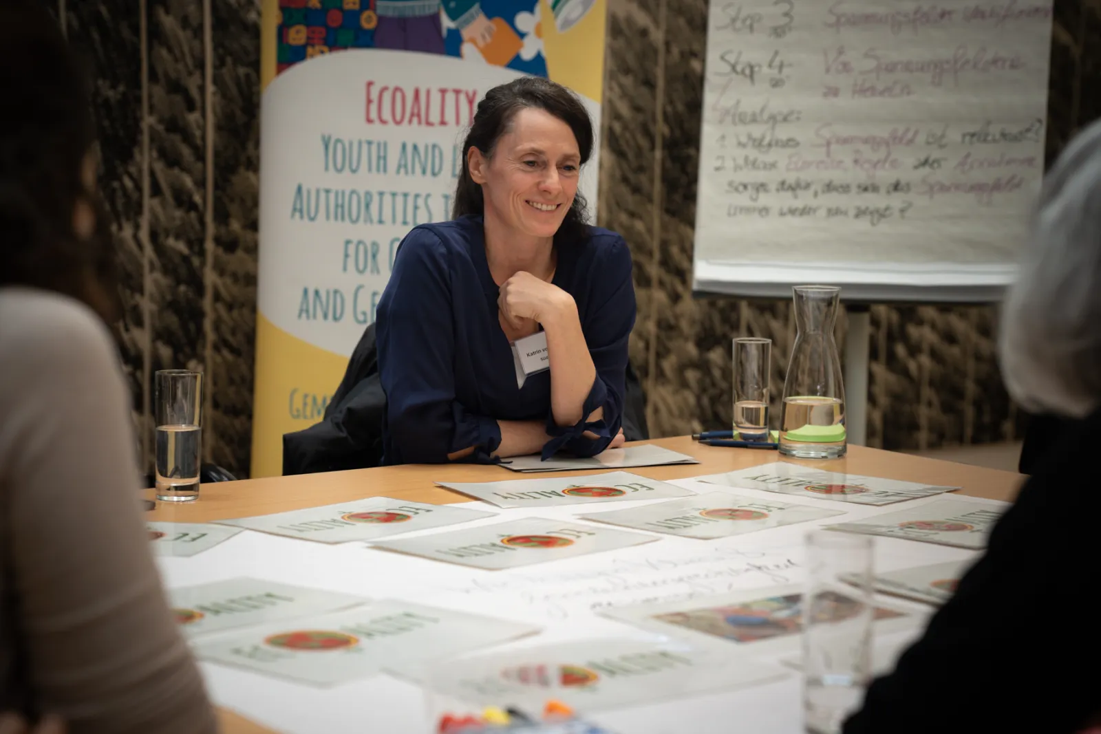 Person sitzt lächelnd an einem Konferenztisch bei einem Workshop, Banner im Hintergrund und Flipchart sichtbar, Unterlagen auf dem Tisch.