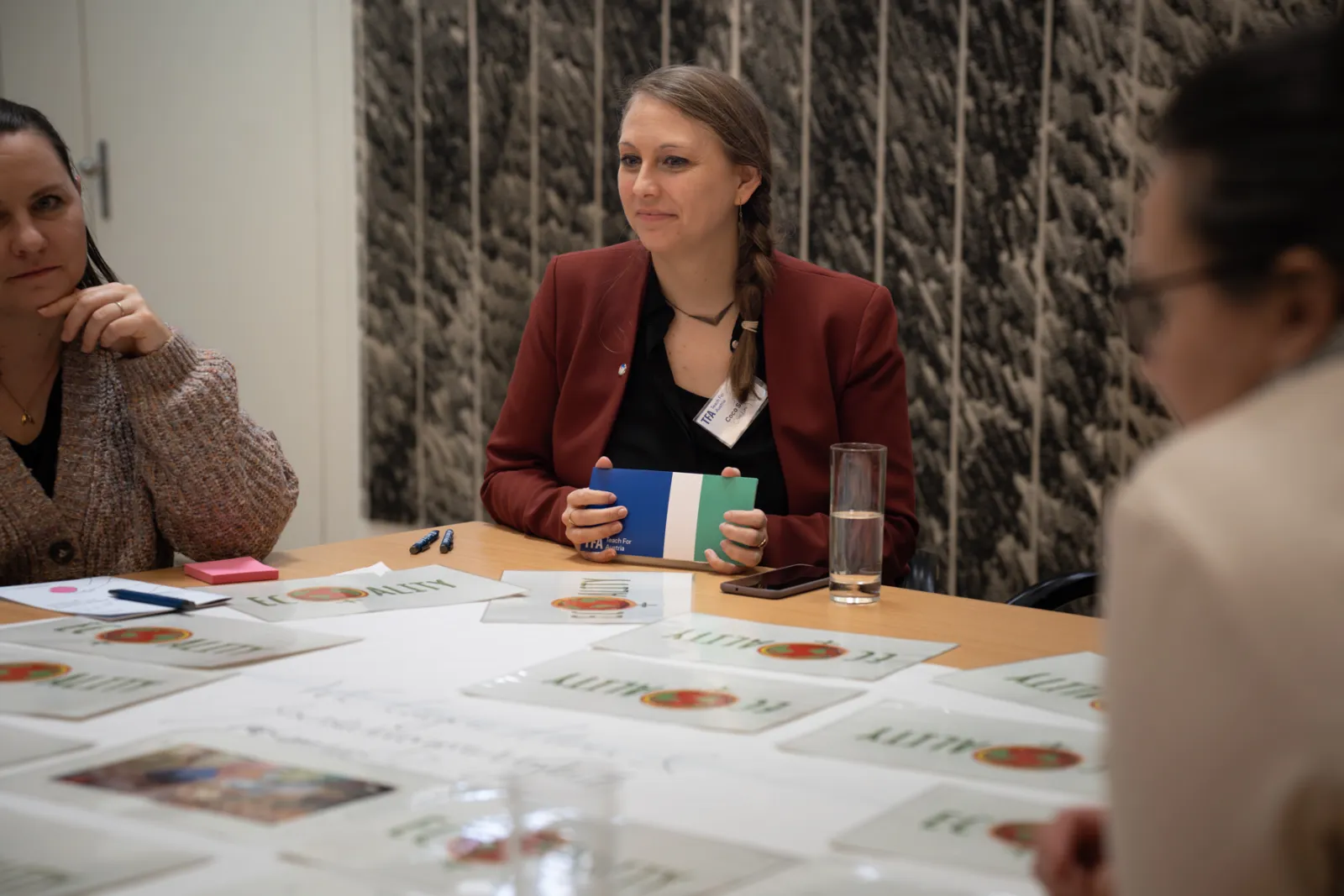 Person im roten Blazer sitzt an Konferenztisch, hält blau-grünes Notizbuch; Tisch mit Unterlagen, weitere Personen im Hintergrund.