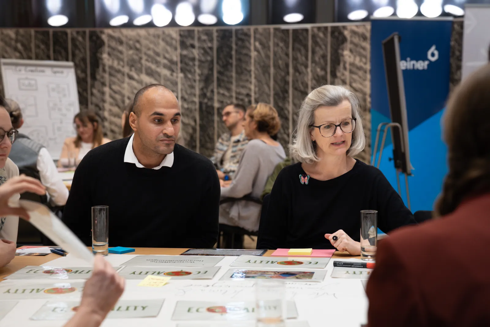 Zwei Personen diskutieren an einem Workshop-Tisch in einem Konferenzraum, umgeben von Unterlagen.