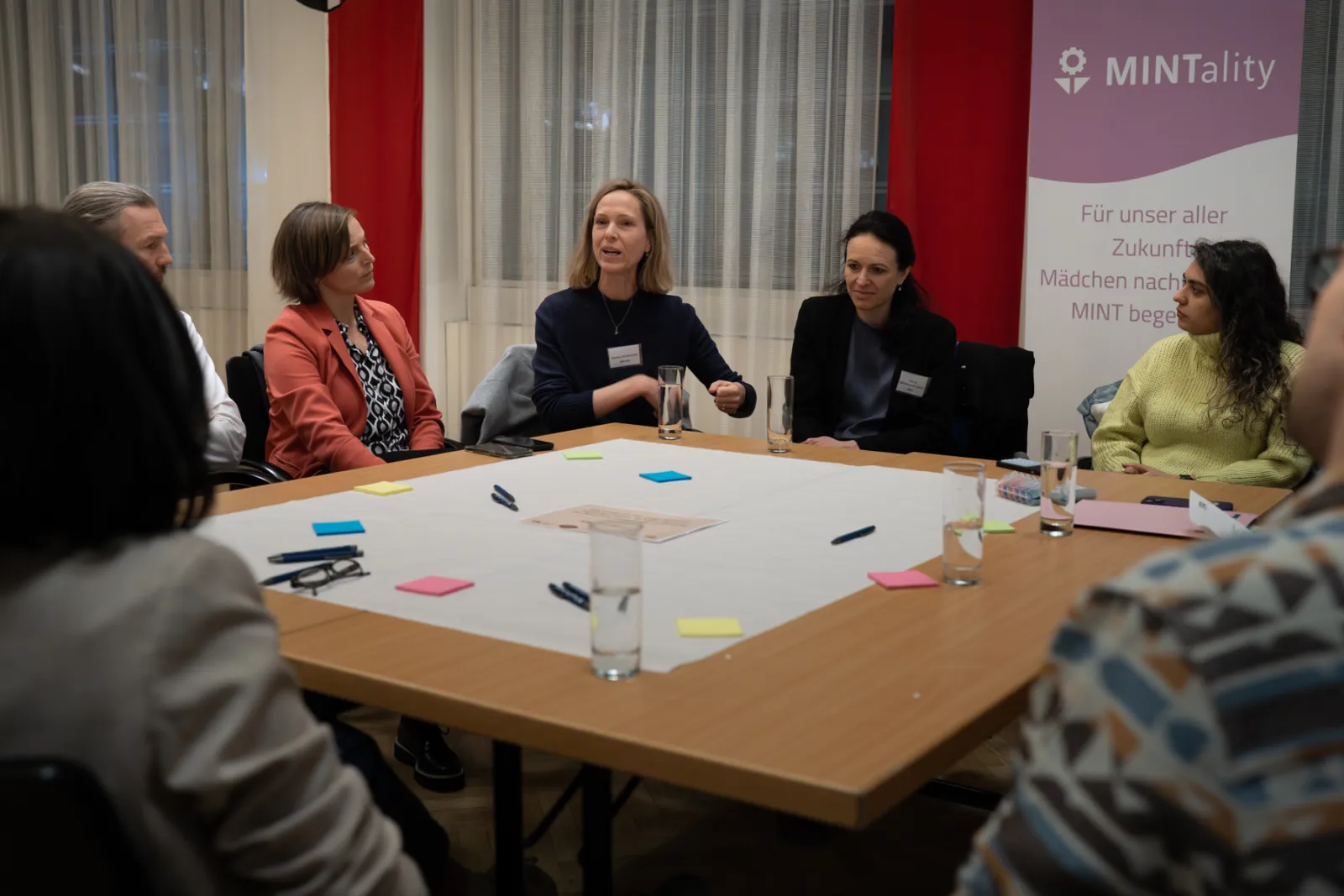 Mehrere Teilnehmende diskutieren an einem runden Konferenztisch; Notizzettel, Stifte; MINTality-Banner im Hintergrund.
