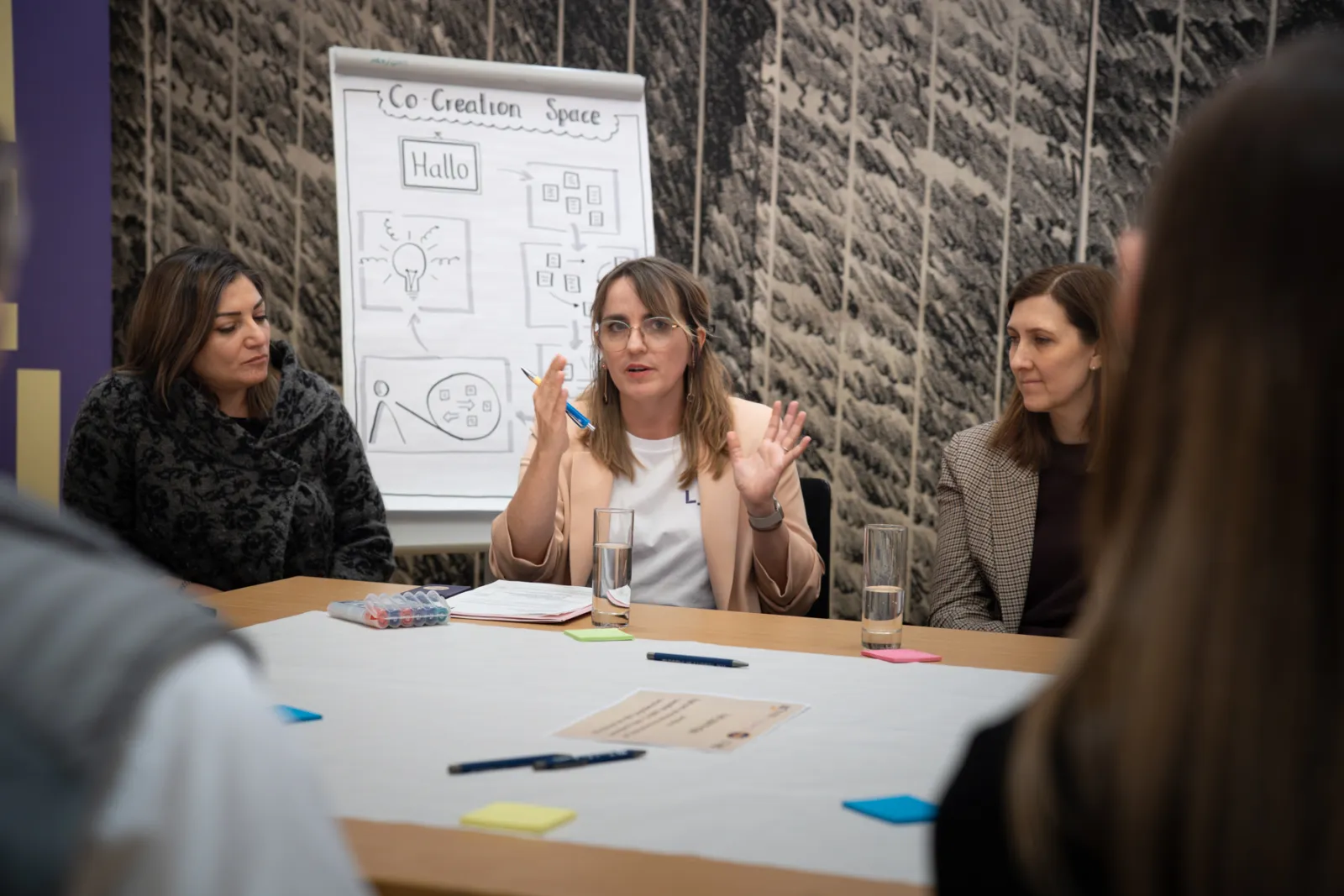 Leitende Person erklärt während Workshop am Tisch vor Whiteboard; zwei weitere Personen hören zu; Schreibmaterialien und Gläser sichtbar.
