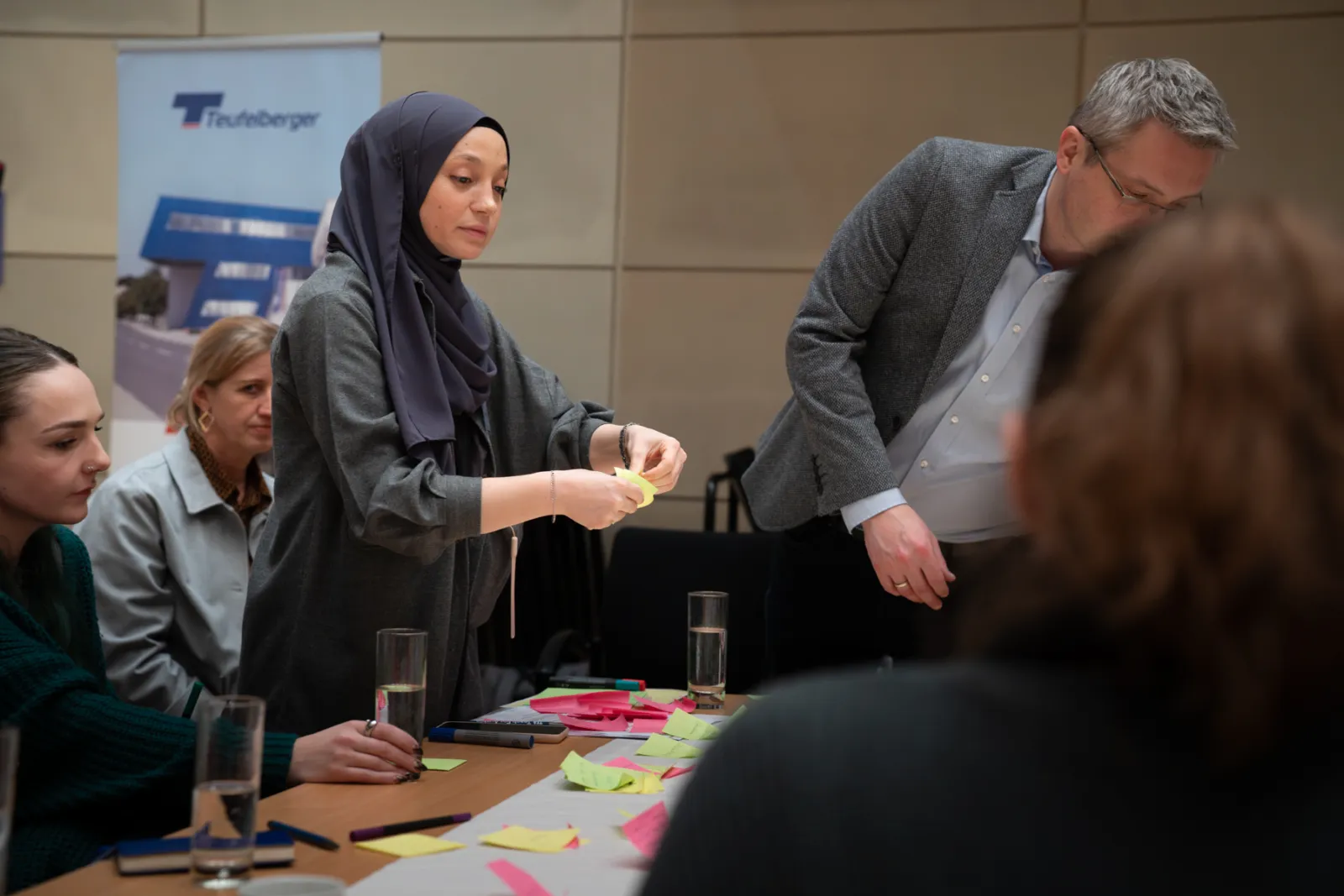 Person mit Hijab ordnet Haftnotizen auf Konferenztisch im Besprechungsraum, während weitere Teilnehmende zuschauen.