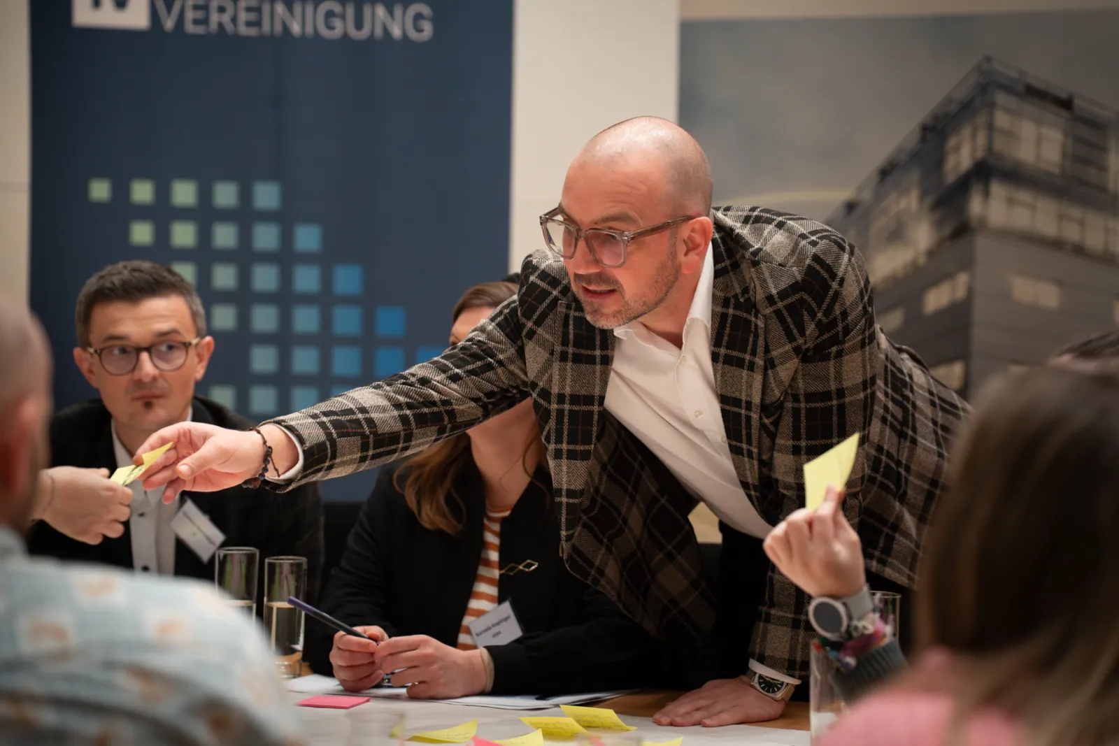 Person mit Glatze und Brille in kariertem Sakko reicht am Tisch Post-it an weitere Person; Banner VEREINIGUNG im Hintergrund