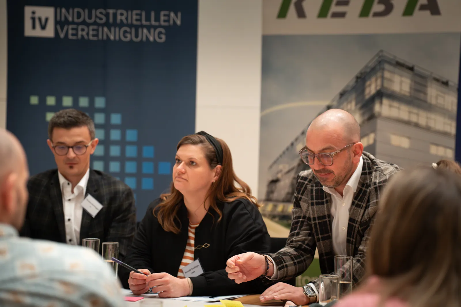 Paneldiskussion mit drei Personen am Konferenztisch; rechts gestikuliert eine Person mit Brille, mittlere notiert, links blickt ins Publikum.