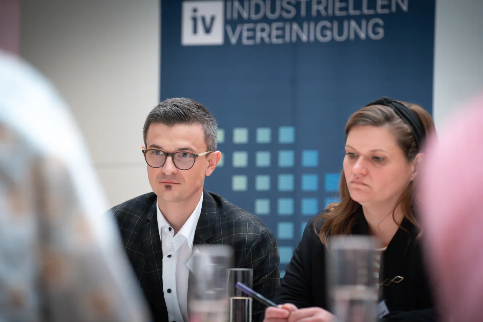 Zwei Personen bei einer Konferenz sitzen am Tisch; Banner der Industrievereinigung im Hintergrund