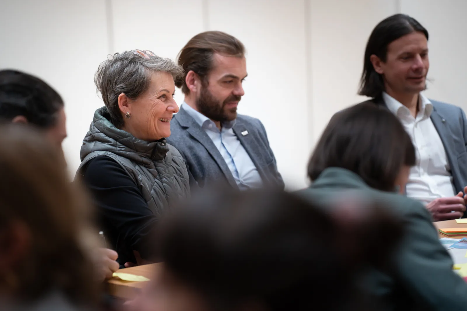 Gruppe von Personen bei einer Besprechung; eine lächelnde ältere Person in Weste sitzt am Tisch, im Hintergrund weitere Teilnehmende.