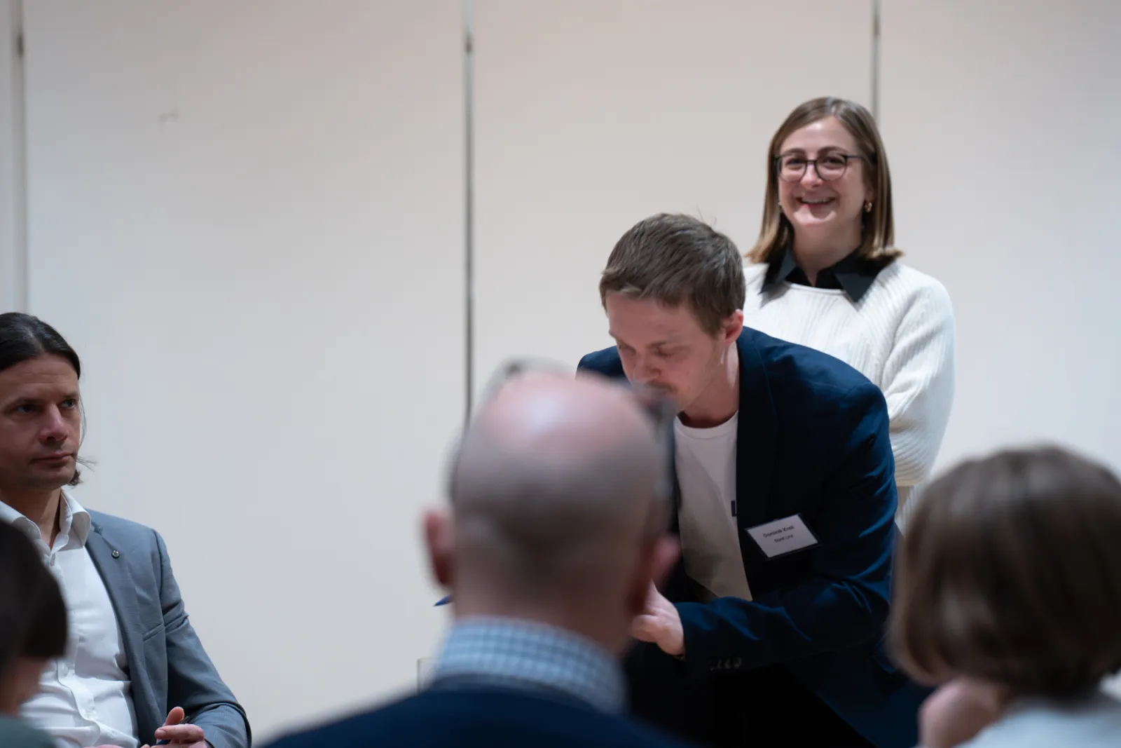 Mann im dunkelblauen Blazer beugt sich vor; dahinter steht eine lächelnde Person in weißem Pullover; Teilnehmende sitzen im Konferenzraum.