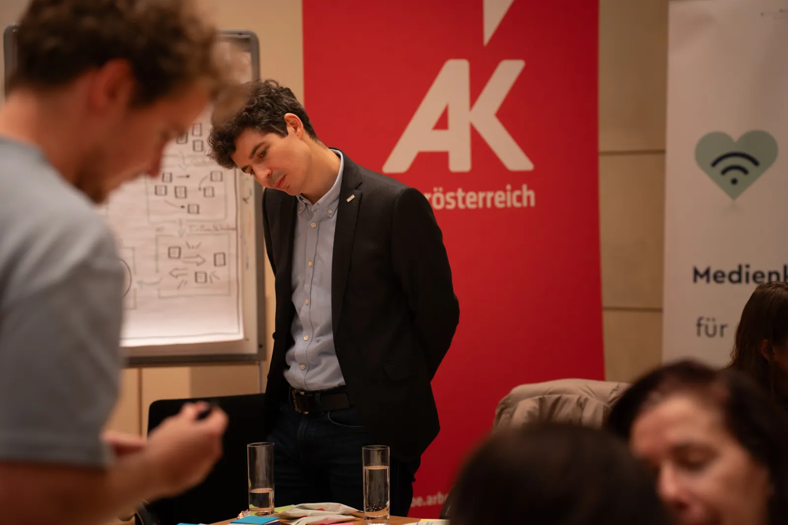 Person im dunklen Blazer beugt sich über ein Whiteboard mit Diagrammen; Konferenzraum, rotes AK-Österreich-Banner im Hintergrund.