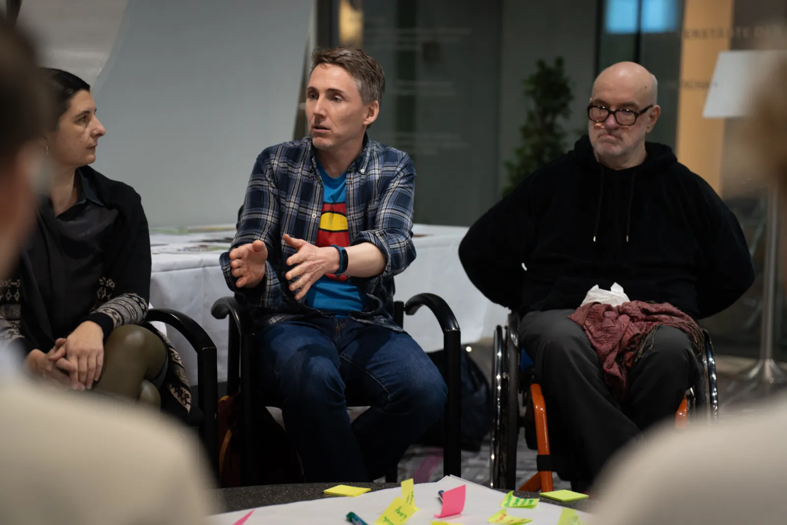 Drei Personen in einer Diskussionsrunde; zentrale Person gestikuliert, daneben Person im Rollstuhl; moderner Innenraum mit Tisch und Post-its.