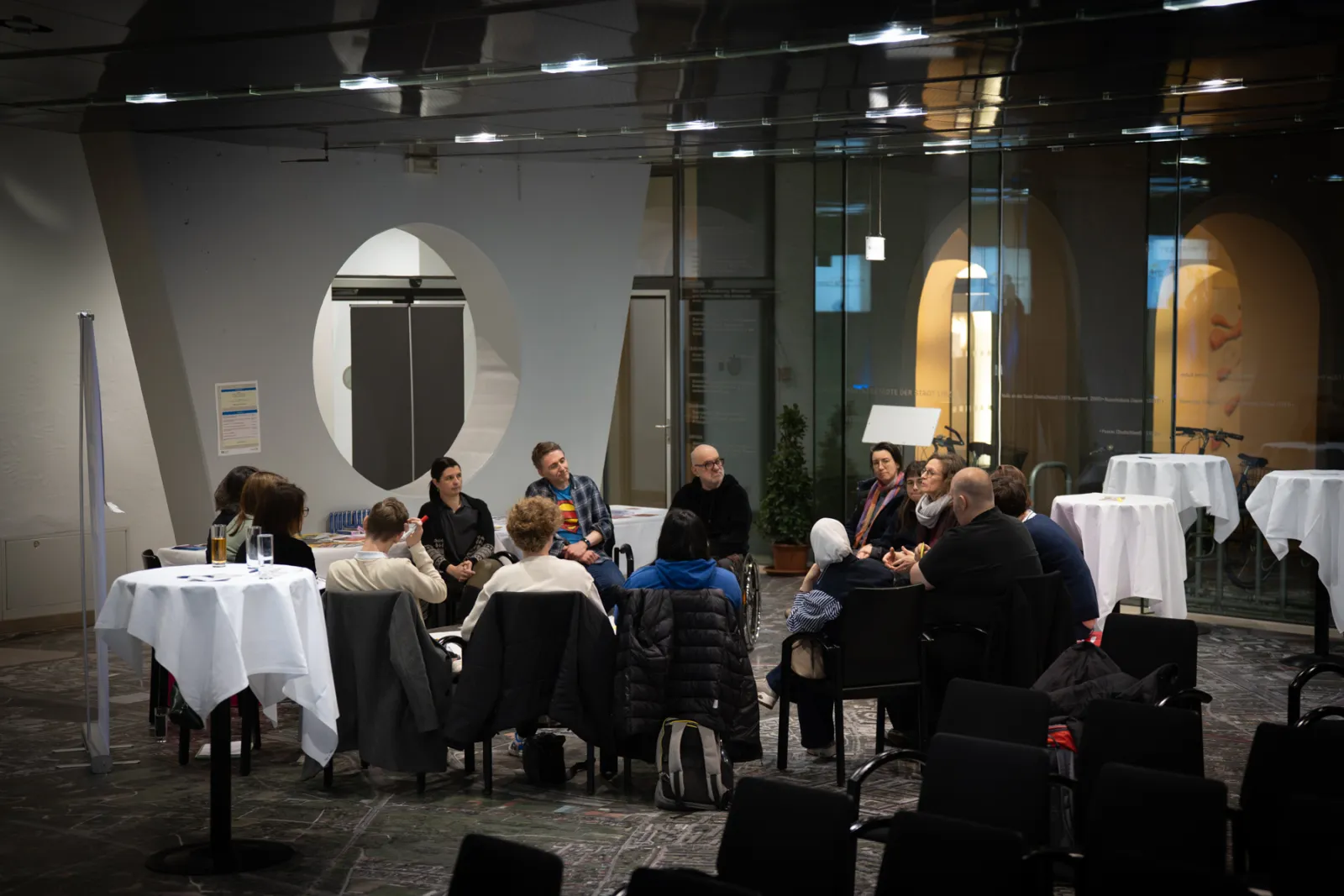 Teilnehmende sitzen kreisförmig in modernem Konferenzraum mit Glasfronten und weißen Tischdecken.