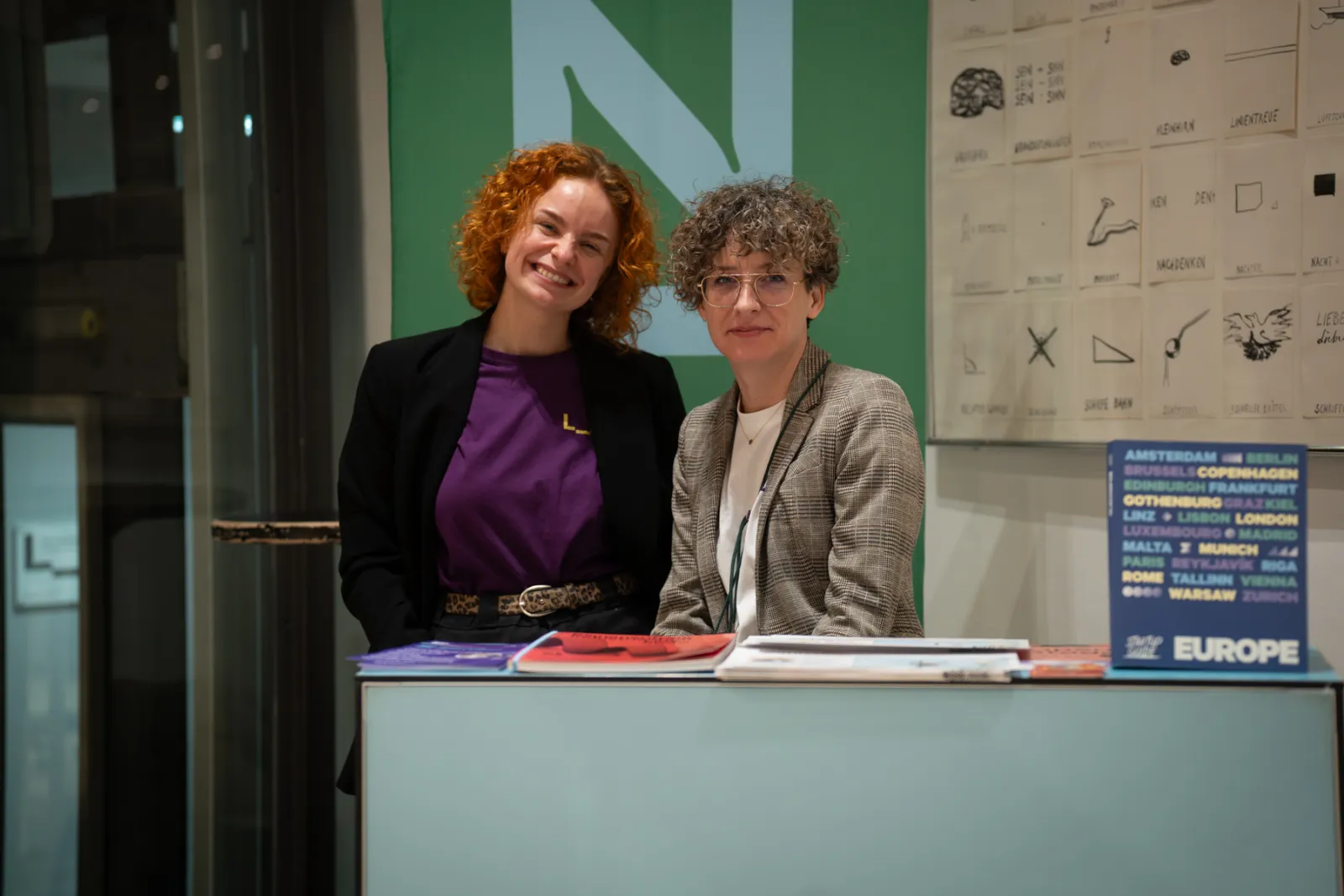 Zwei Personen stehen hinter einem Messestand, lächelnd; Broschüren liegen auf der Theke, grüne Wand im Hintergrund.