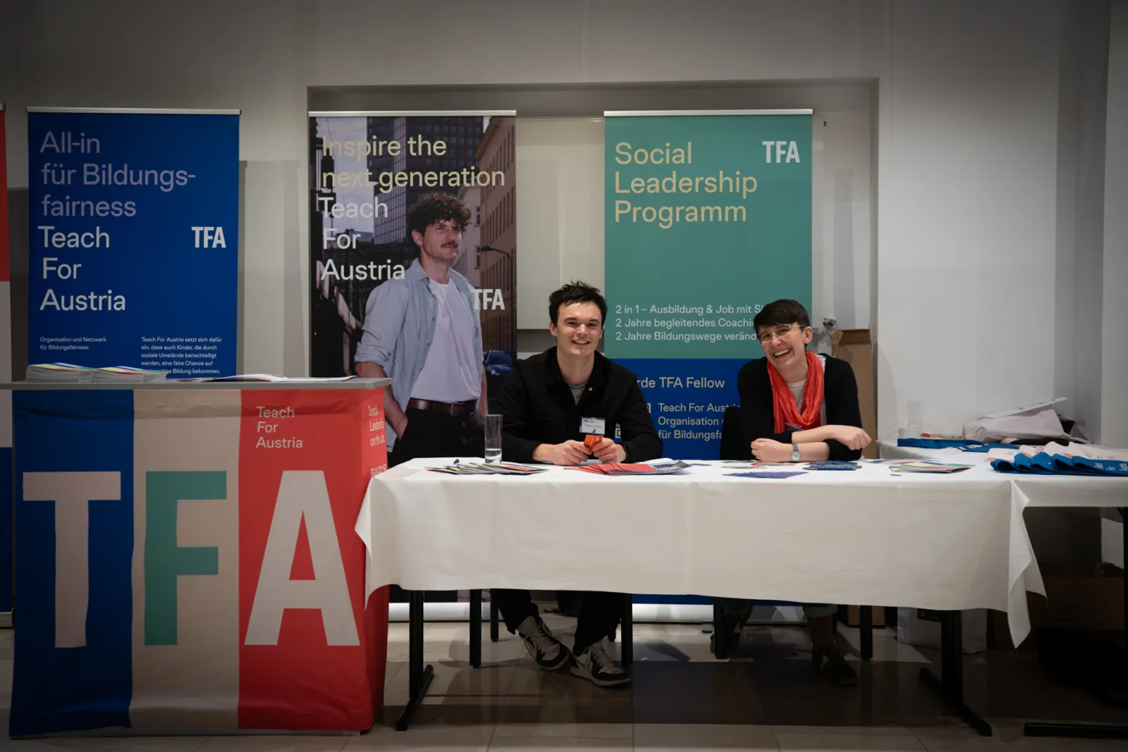 Zwei Personen sitzen hinter einem TFA-Infostand mit Bannern und Materialien bei einer Bildungsmesse.