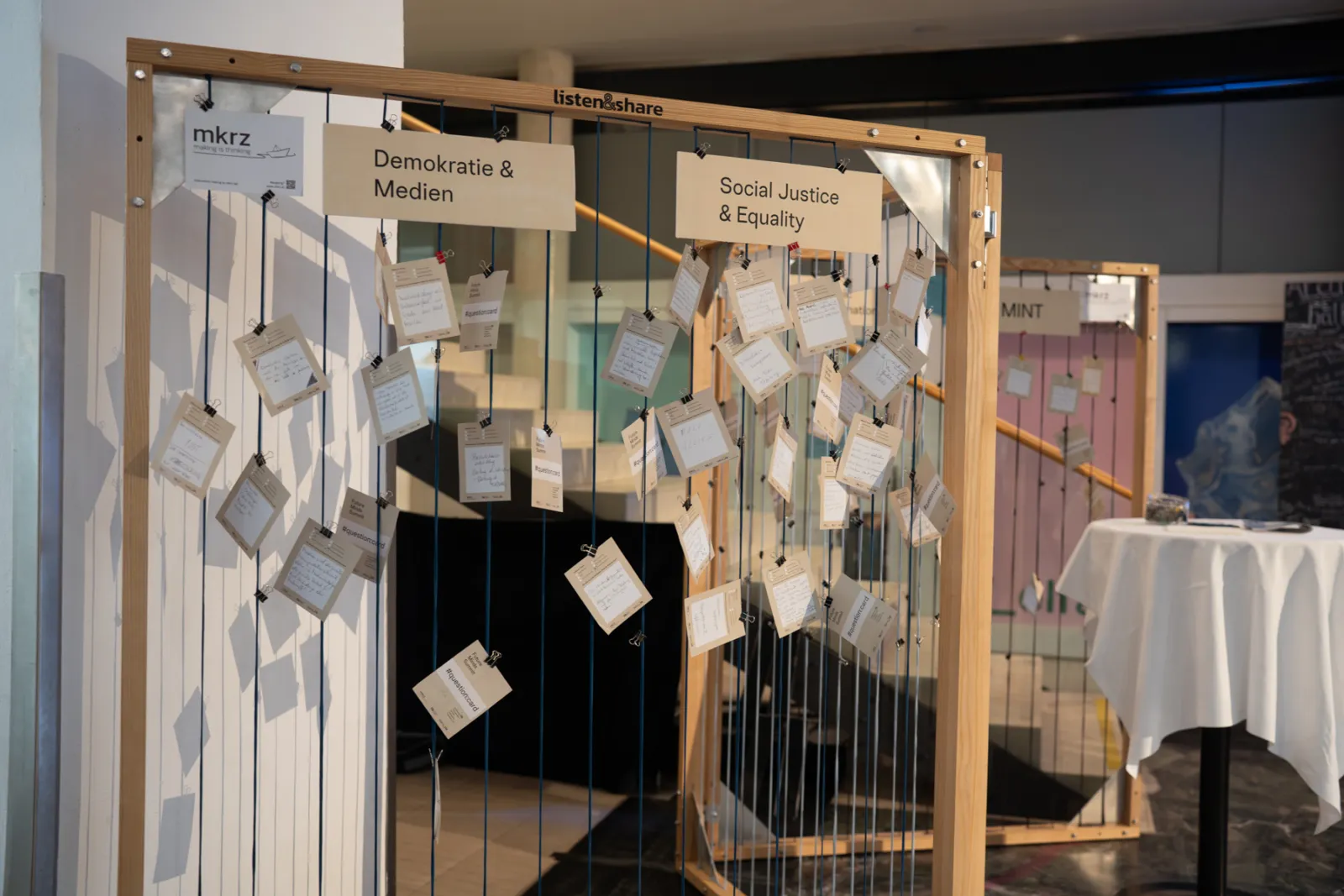 Hängende Zettel an Holzkonstruktion mit Schnüren; Beschilderung 'Demokratie & Medien' und 'Social Justice & Equality' im Ausstellungsraum.