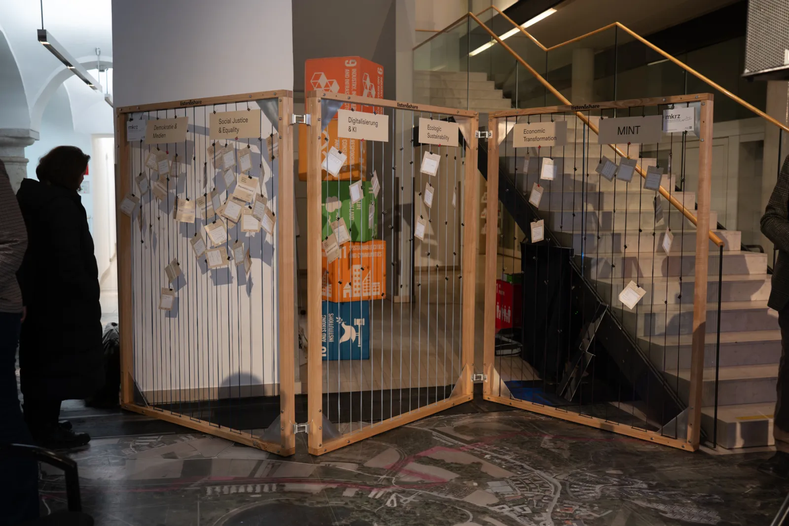 Interaktive Notiz-Installation aus Holzrahmen mit Zetteln an Schnüren, Innenraum-Ausstellung nahe Treppe; Besucher hängen Zettel auf.