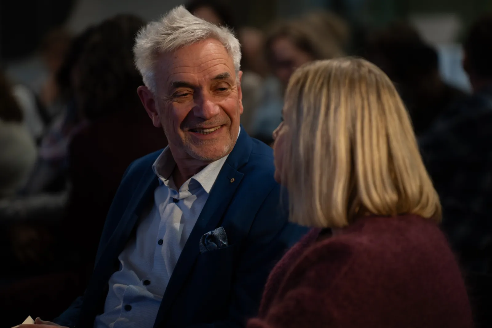 Mann im blauen Anzug lächelt einer Frau zu; beide im Gespräch bei einer Veranstaltung.