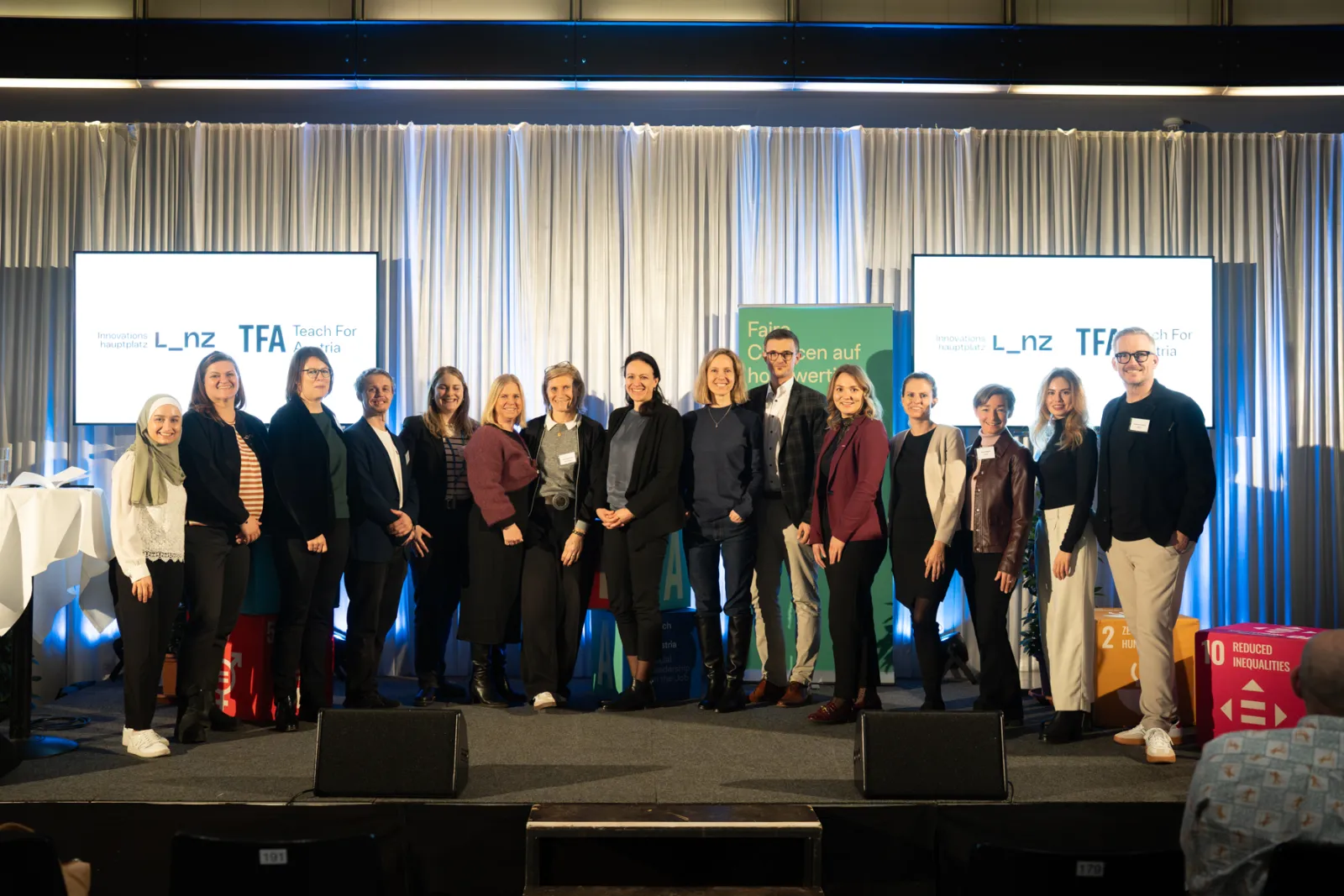 Gruppe von etwa 15 Menschen posiert auf Bühne bei Konferenz; Vorhänge, zwei Bildschirme mit Logos im Hintergrund.