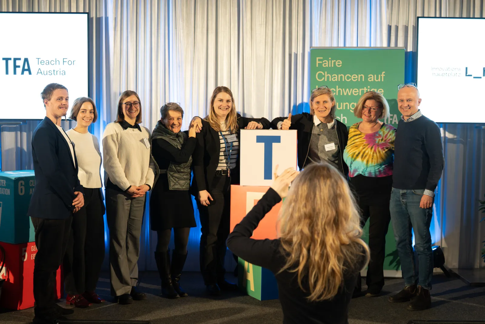 Neun Personen posieren auf einer Bühne hinter bunten Buchstabenwürfeln; im Vordergrund fotografiert eine Person das Gruppenbild.