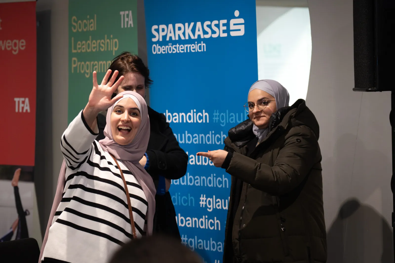 Zwei Frauen mit Hijab bei einer Veranstaltung; eine winkt in die Kamera, die andere zeigt auf sie, vor bunten Bannern.