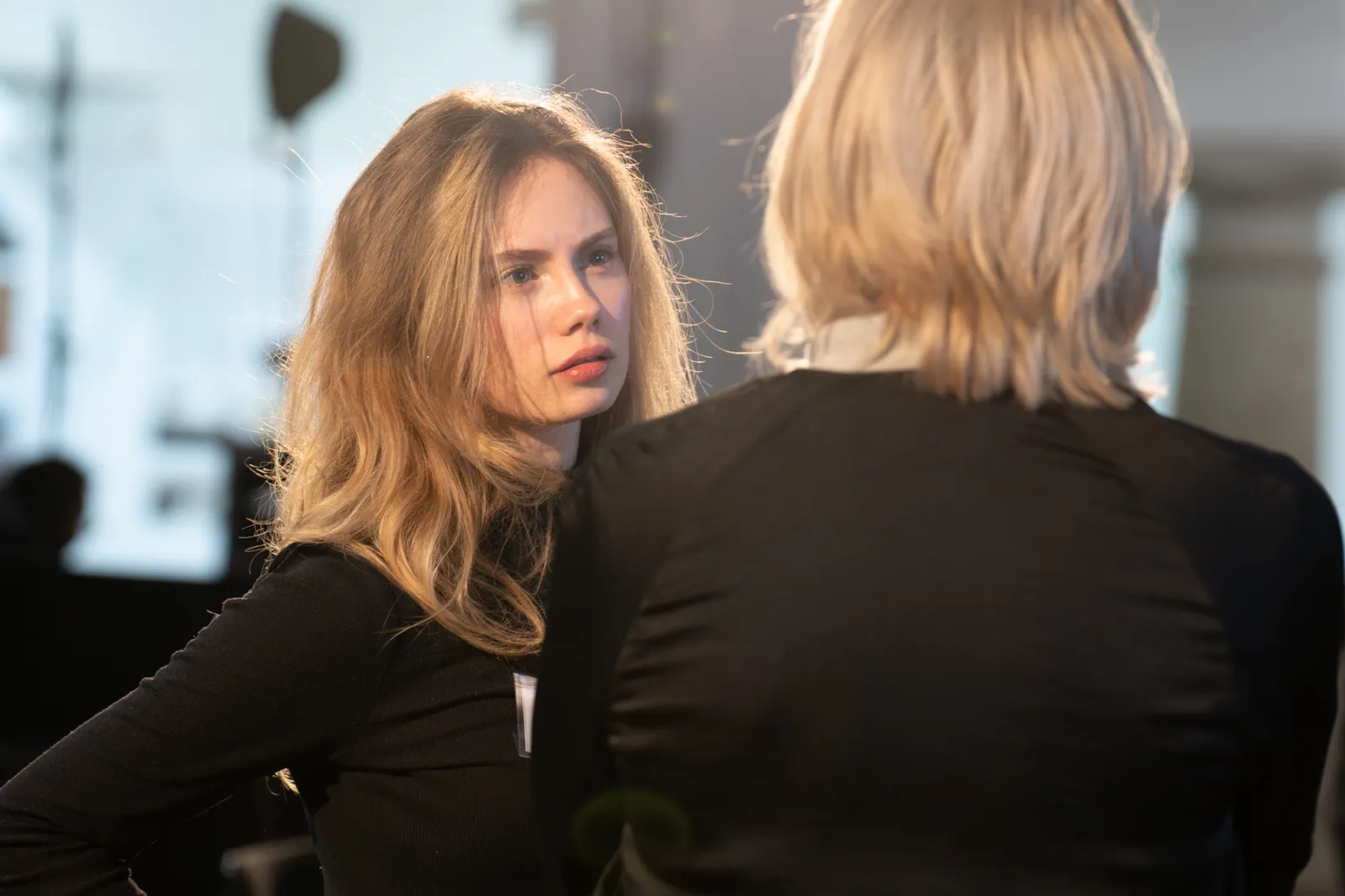 Blonde Frau in schwarzem Oberteil schaut eine andere Person von hinten an; Studio-Umgebung im Hintergrund.