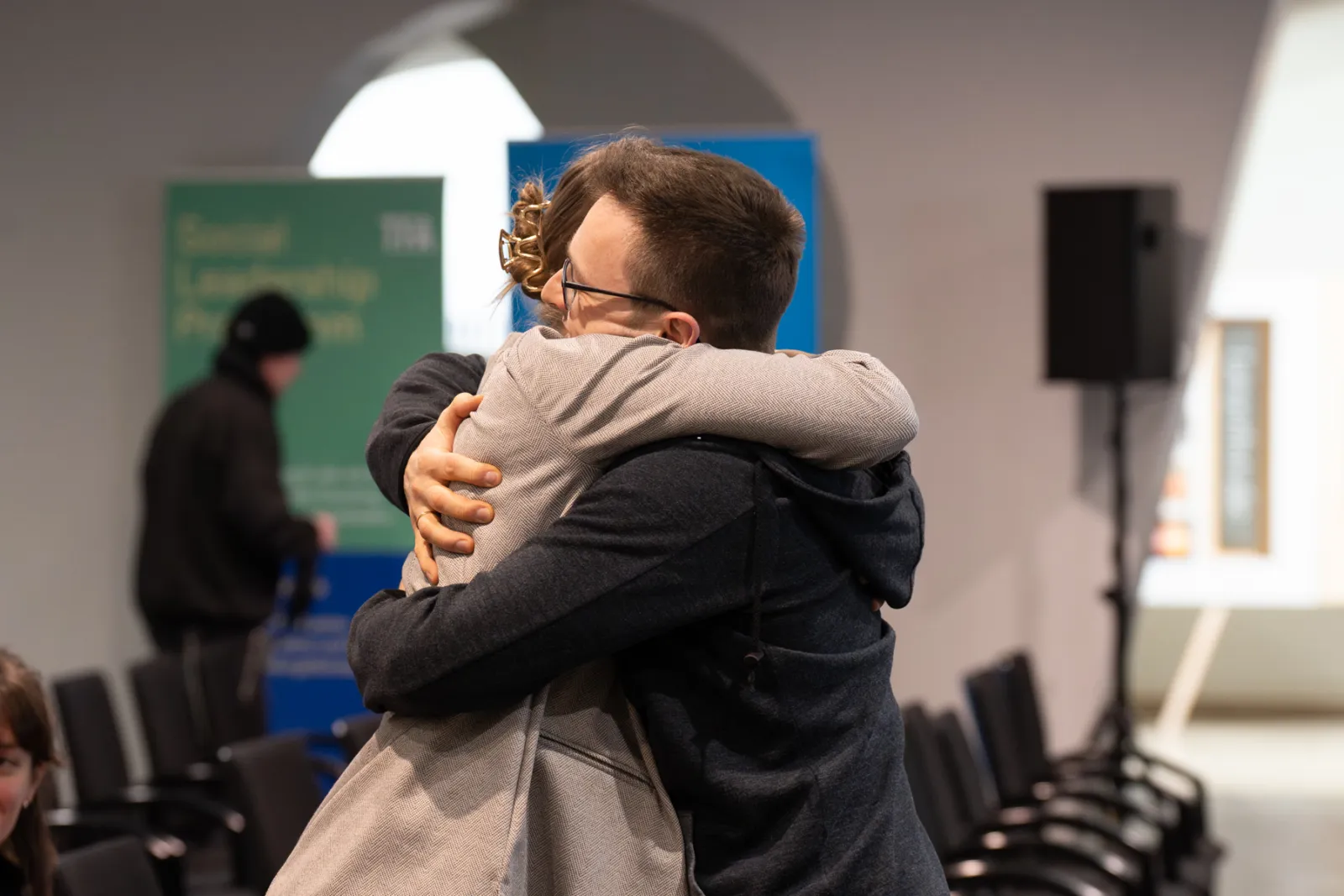 Zwei Personen umarmen sich in einem Konferenzraum, im Hintergrund Stühle und Banner.