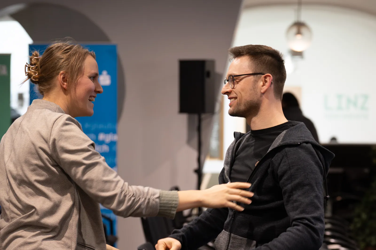 Zwei Personen begrüßen sich lächelnd in einem Veranstaltungsraum; eine Person reicht der anderen die Hand.