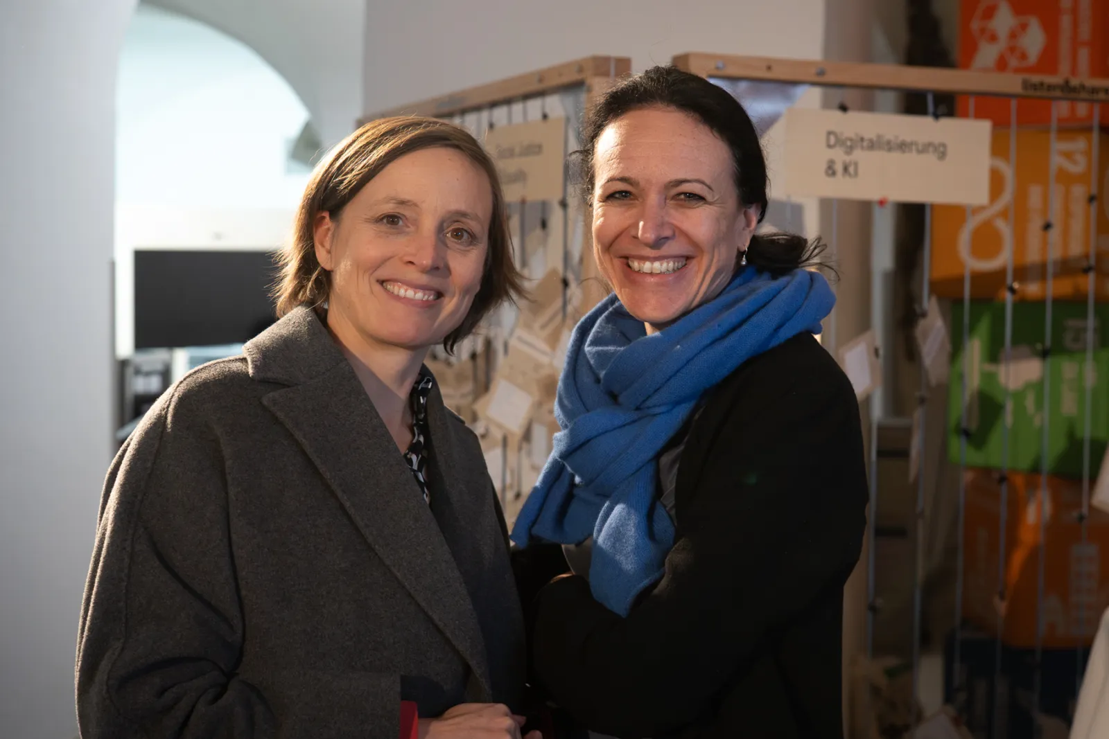 Zwei Personen lächeln in einem Ausstellungsraum; hinter ihnen Stände mit Digitalisierung & KI.