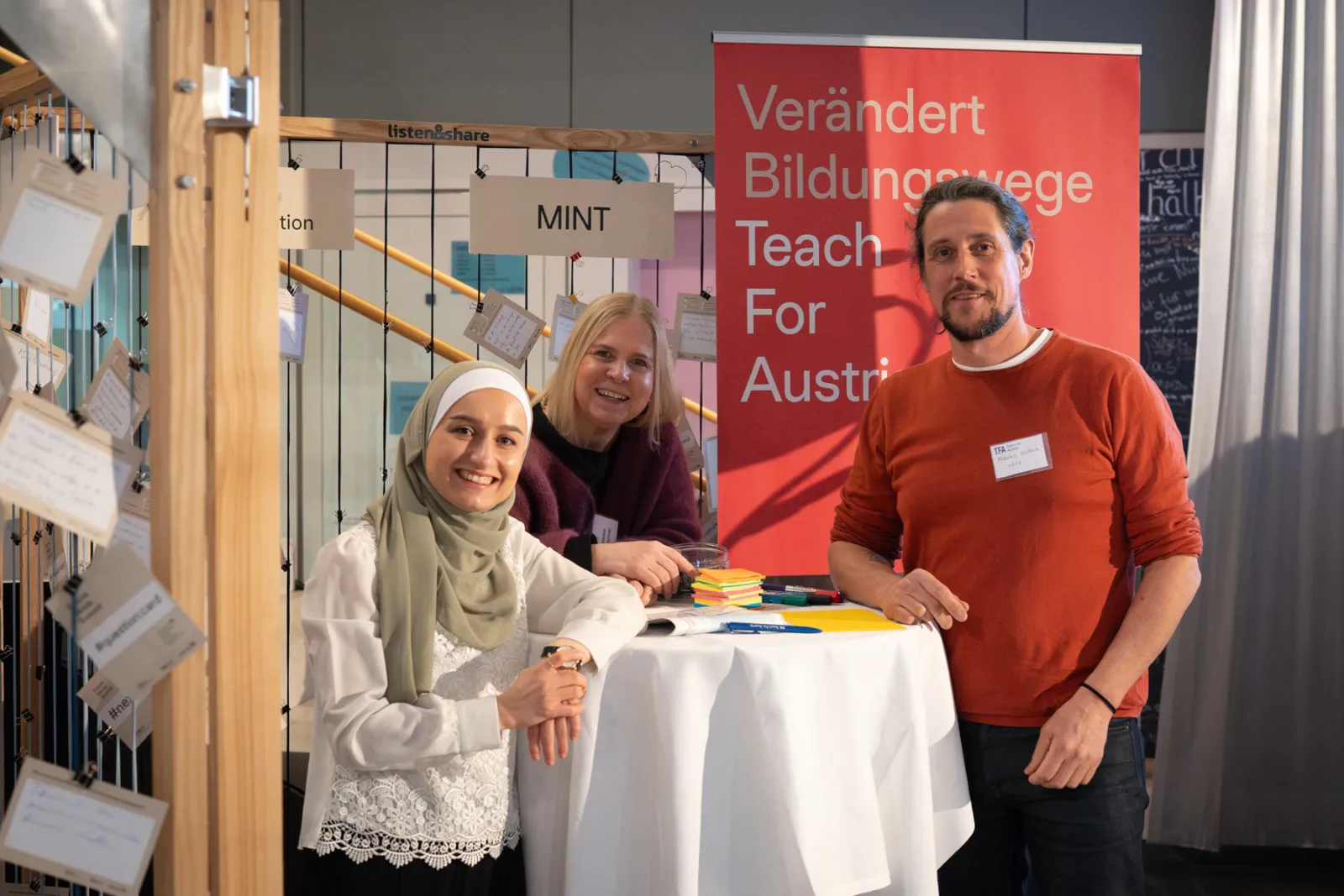 Drei Menschen stehen an einem runden Stehtisch beim Messestand von Teach For Austria; roter Banner Verändert Bildungswege, Flyer-Stapel.