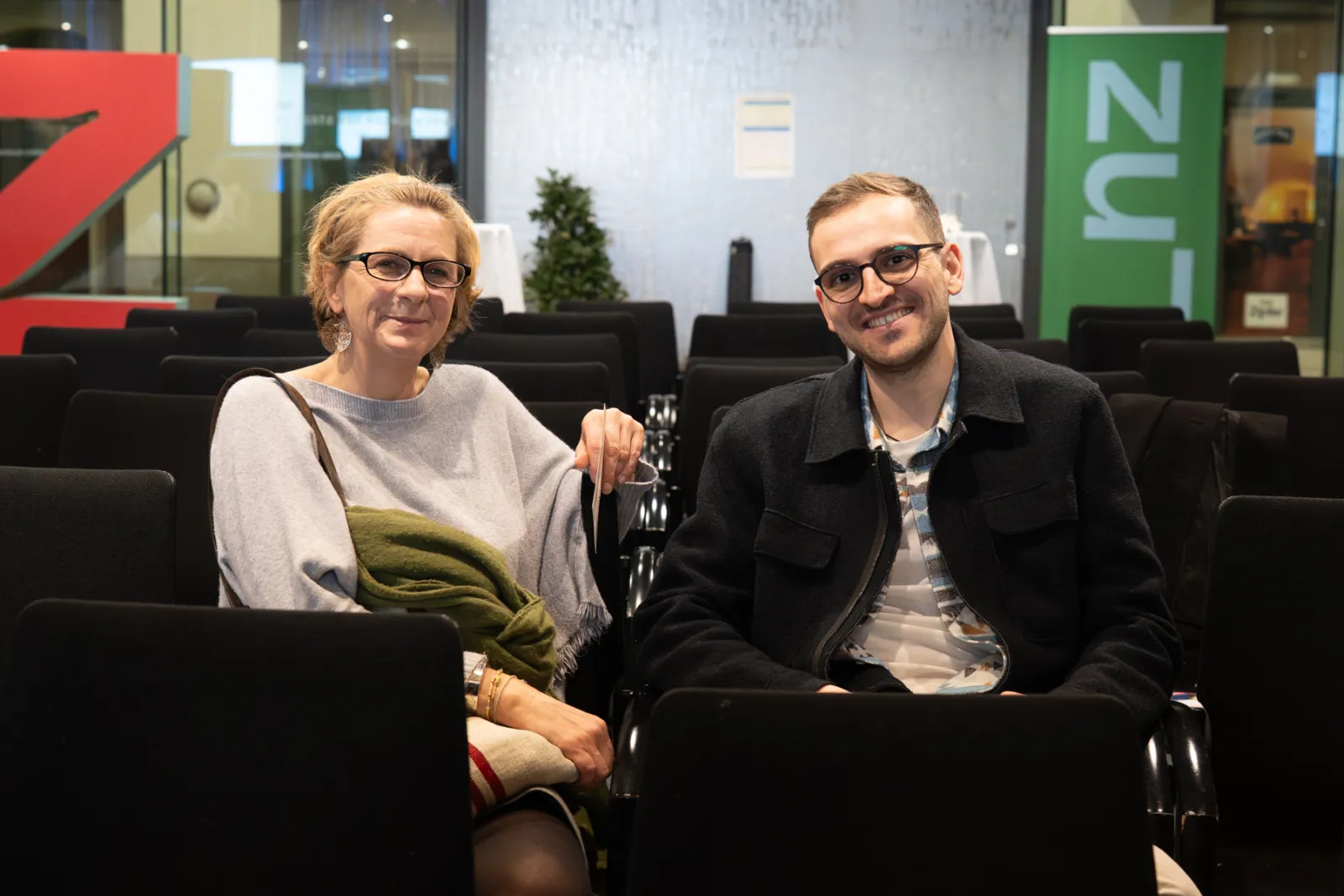 Zwei Personen sitzen in schwarzen Sitzreihen eines Konferenzsaals und lächeln in die Kamera.